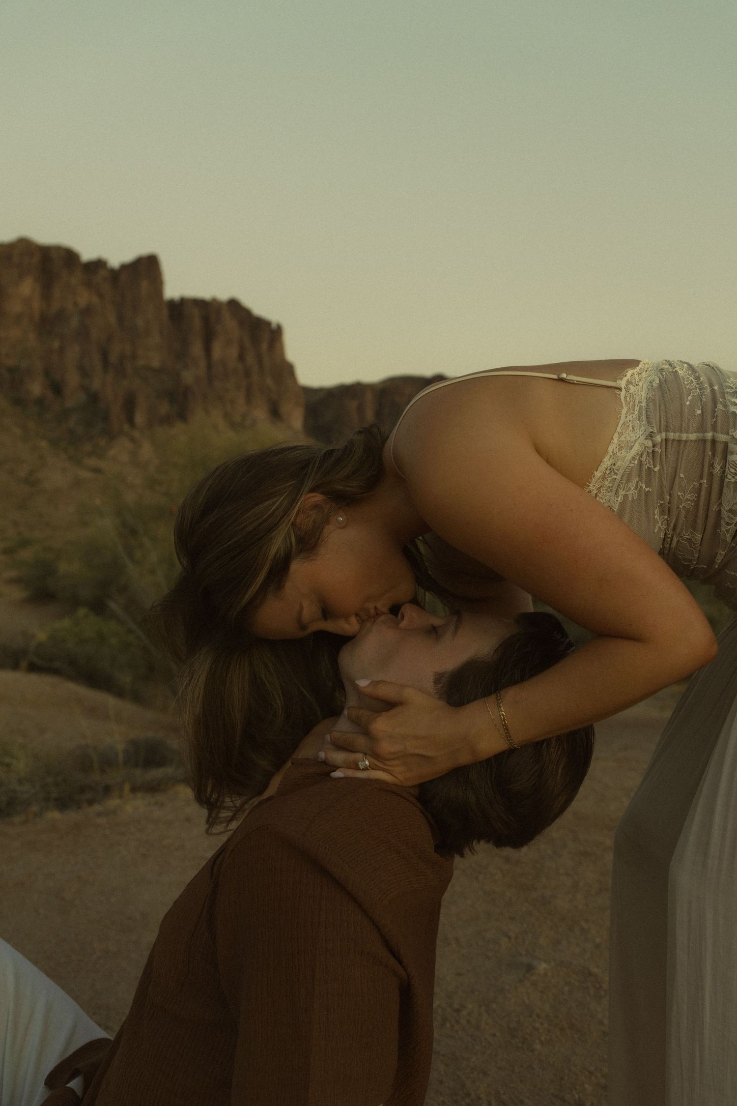 Two women share a romantic kiss at golden hour with desert mountains and cacti in the background.
