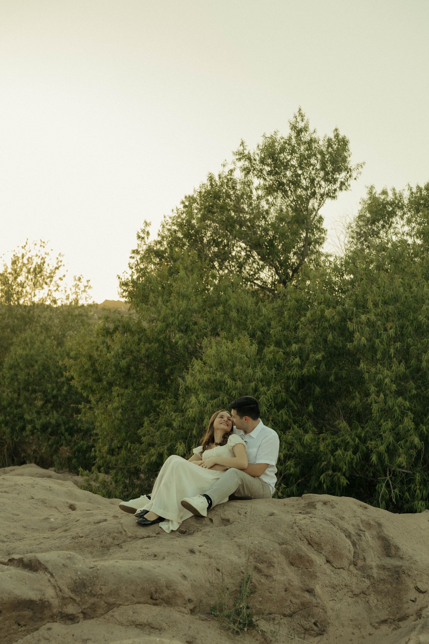 Romantic couple sitting together on sandy rocks surrounded by green trees at dusk.