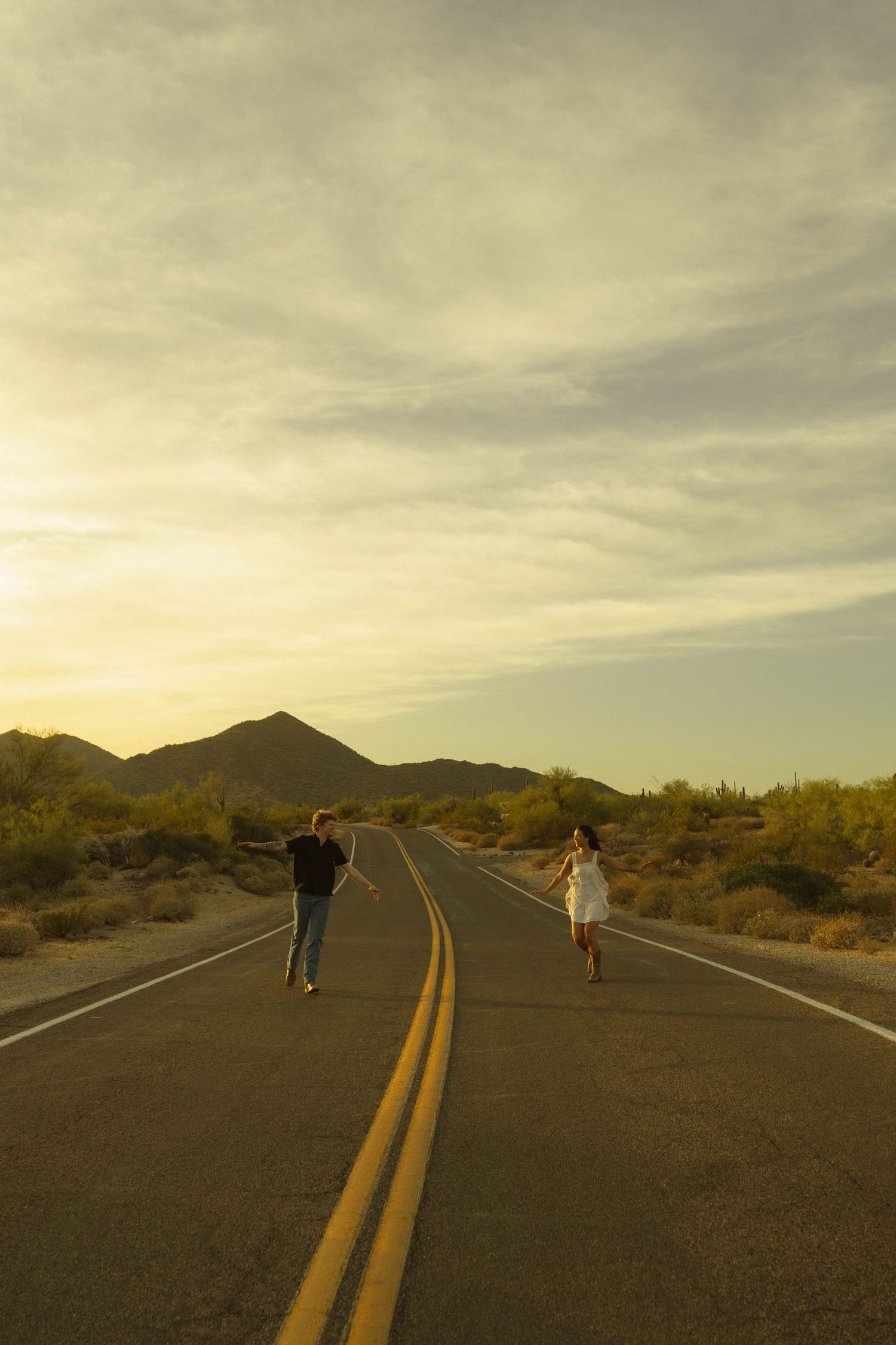 Couple walking toward each other on an empty desert road at sunset with mountains and cacti in background.