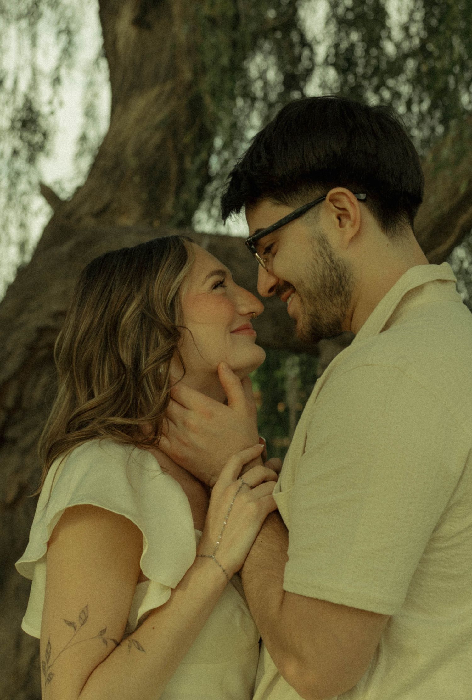 A couple shares a romantic moment under a tree, the man gently holding the woman's face as they smile at each other.