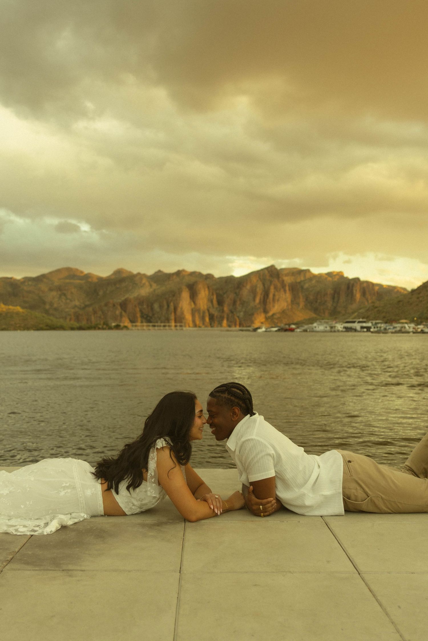 Couple in white lying face-to-face on a dock by a scenic mountain lake at golden hour.