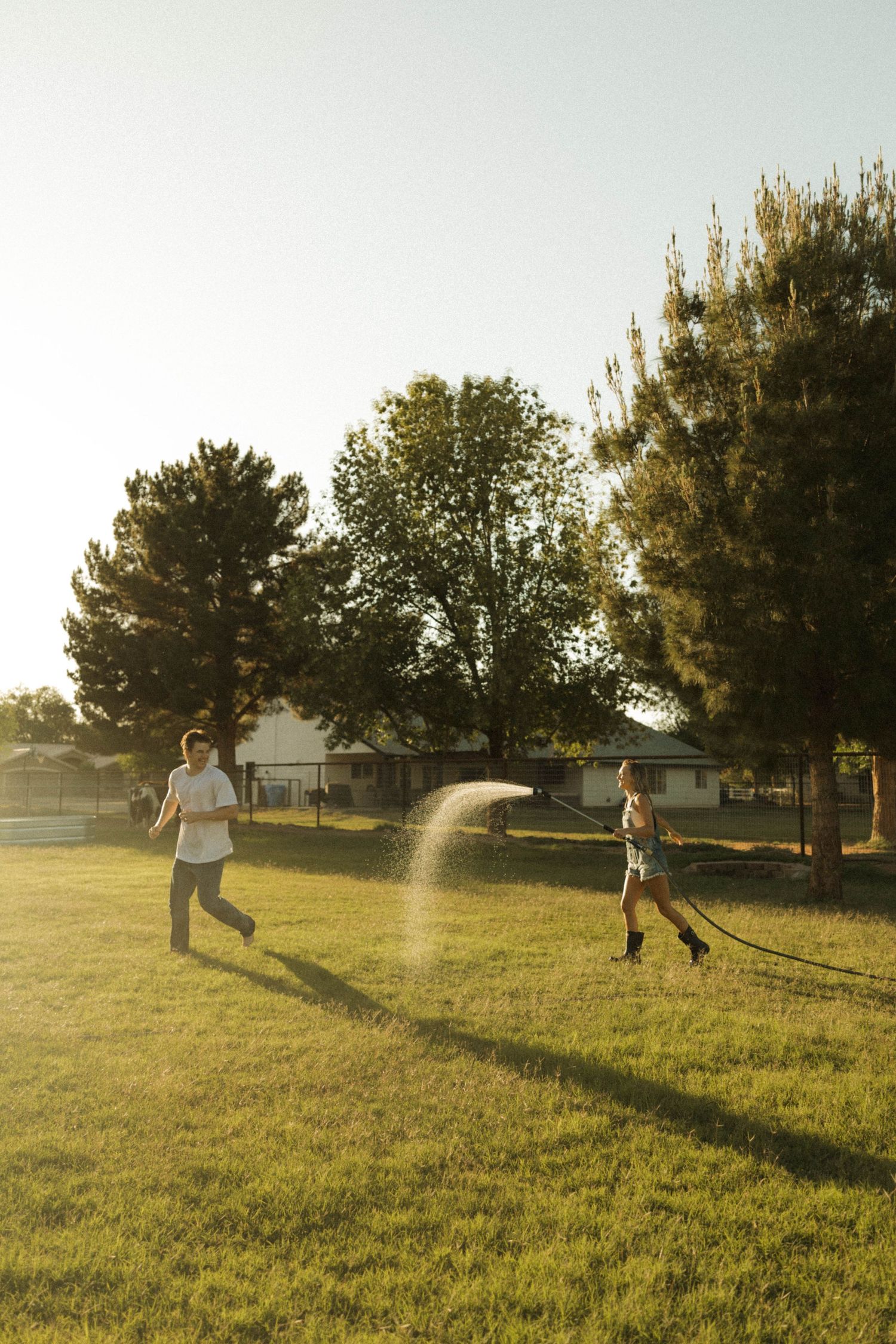 Playful couple chasing each other with a garden hose in a green backyard during golden hour sunset.