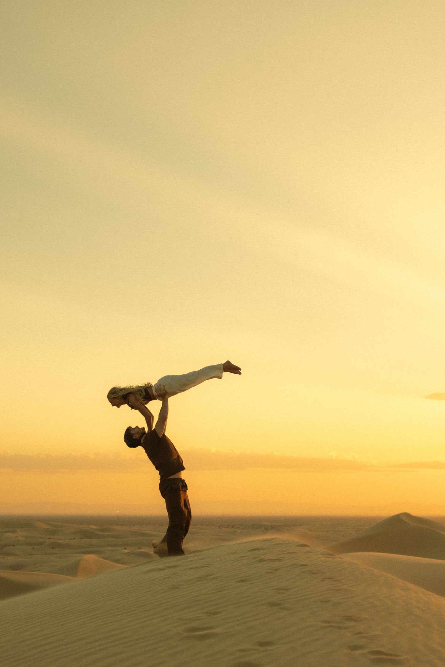 Two acrobats perform a balance act on golden sand dunes at sunset in the desert.