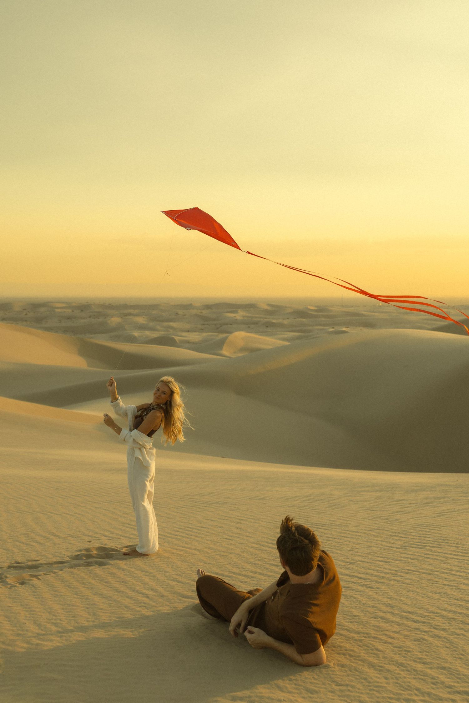 Woman in white outfit flying a red kite on vast sand dunes at golden sunset while man sits nearby.