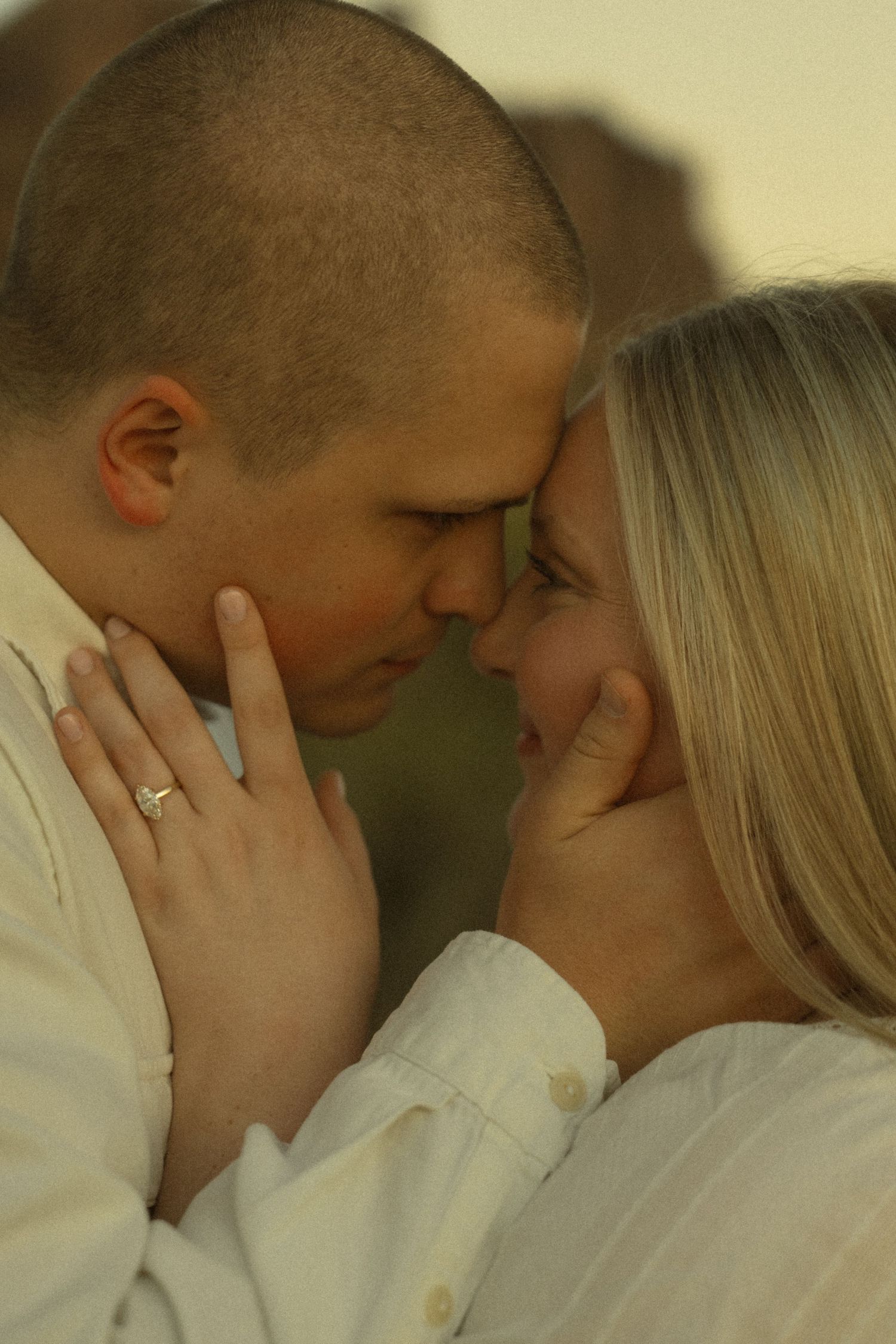 A couple in white shirts share a romantic forehead-to-forehead moment, with the woman wearing a gold ring.
