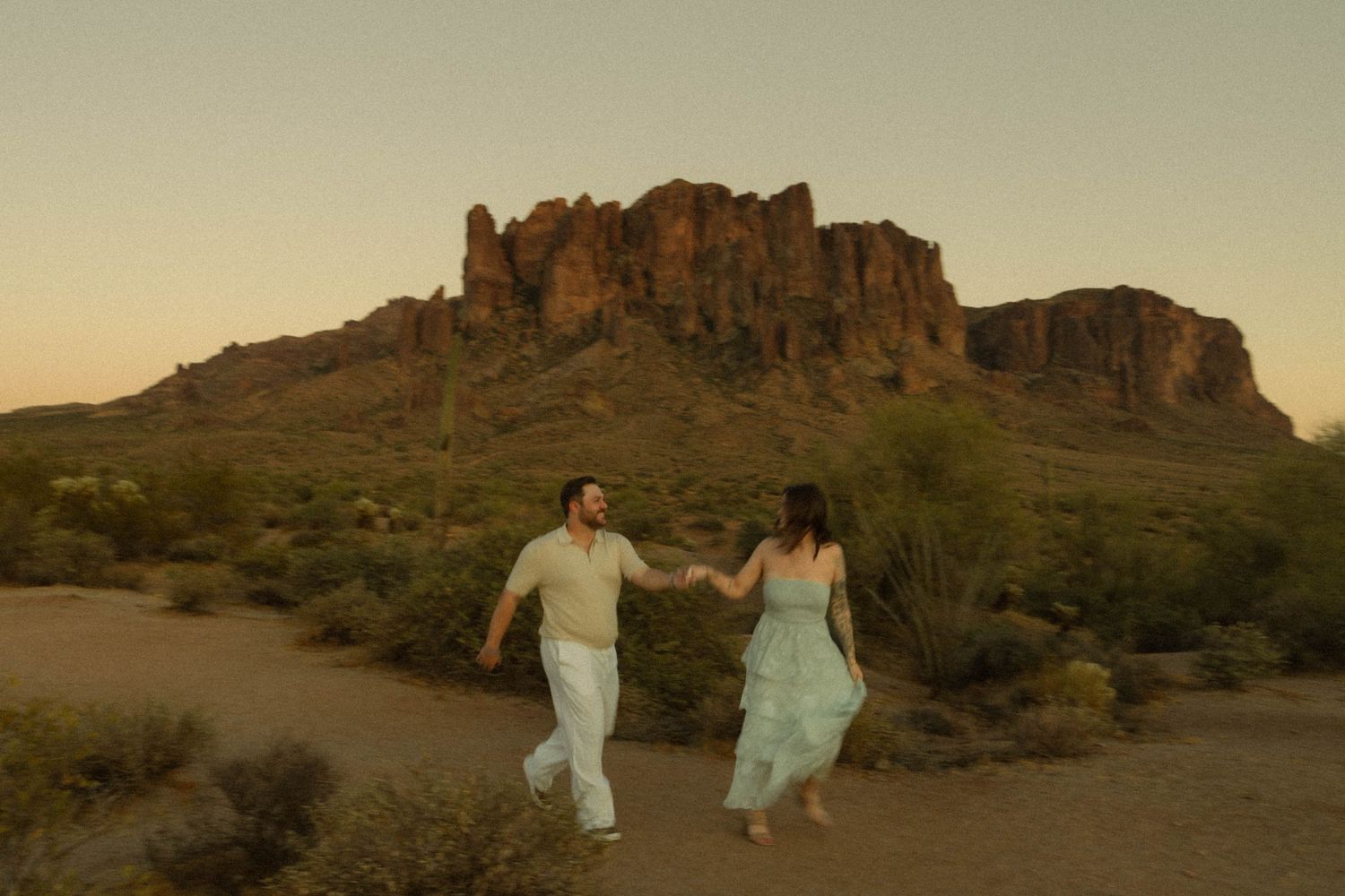 Couple holding hands running through Arizona desert at sunset with Superstition Mountains in background.