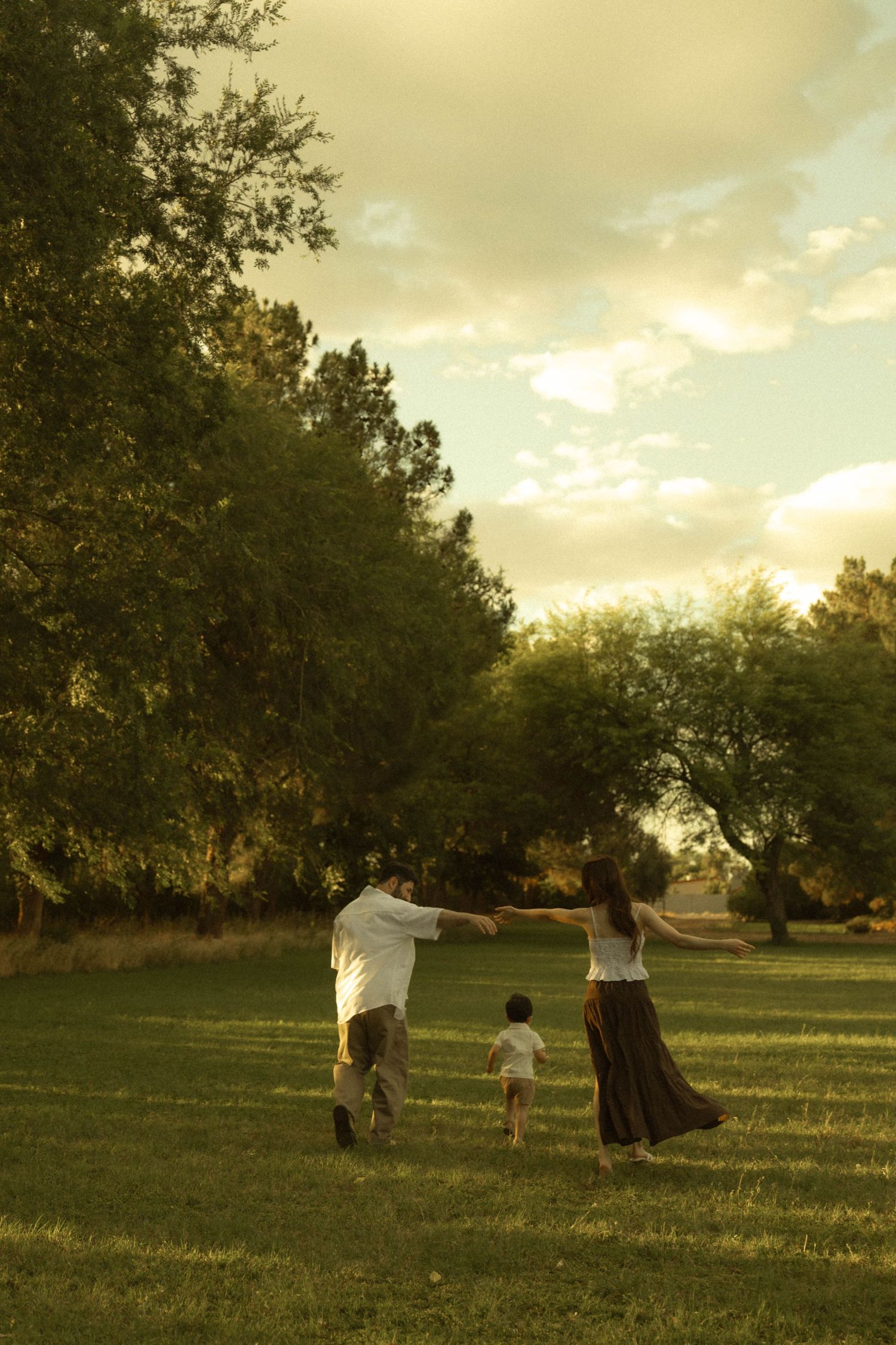Family with young child holding hands walking through a lush green park at golden sunset hour.