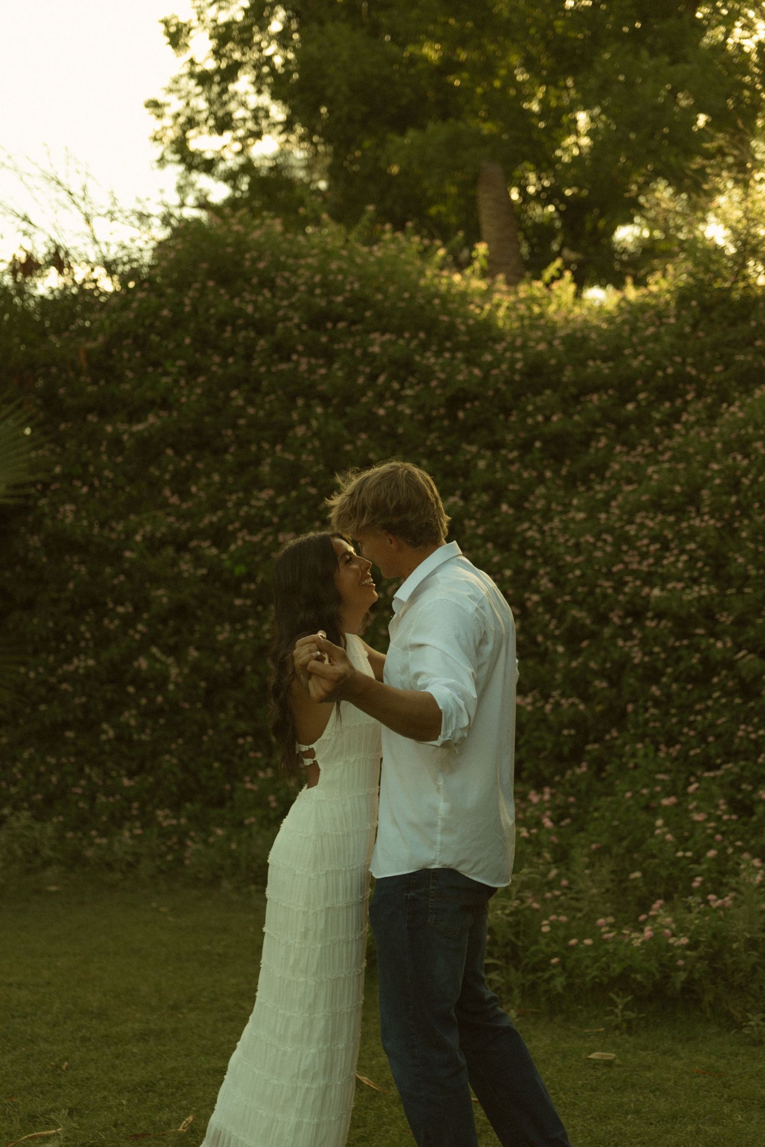 Romantic couple dancing and nearly kissing in a golden-hour garden, woman in white ruffled dress holding hands with man.
