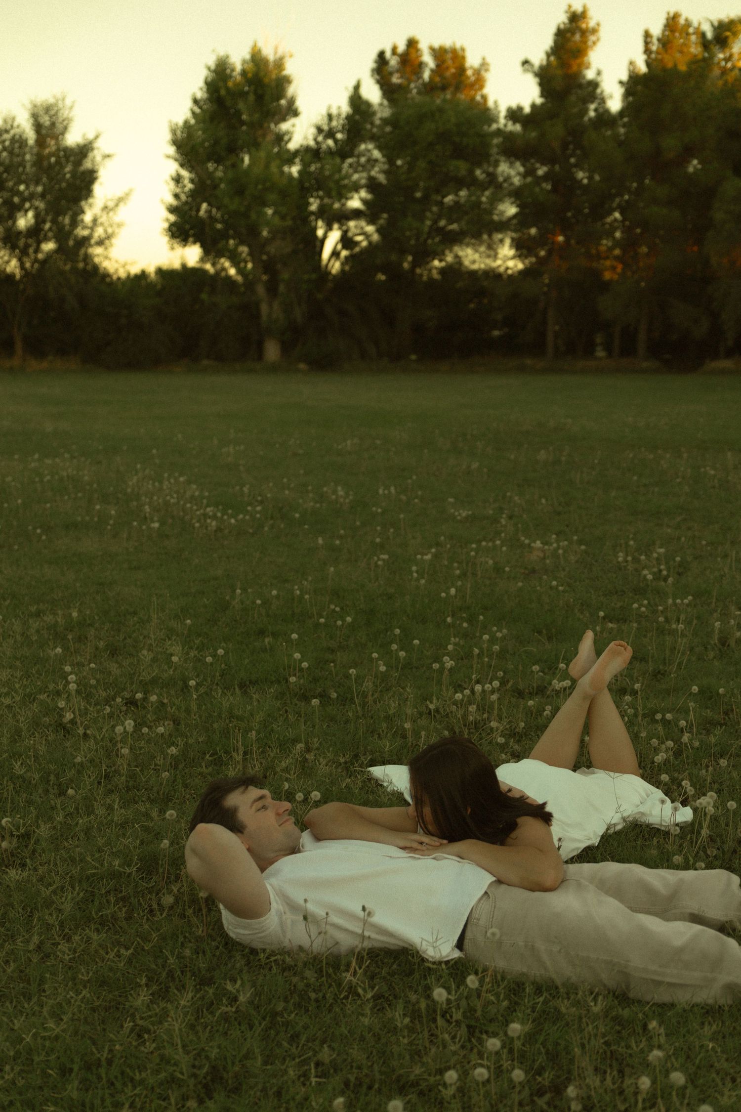 Couple lying together on green grass in a park, relaxing at dusk with trees in background.