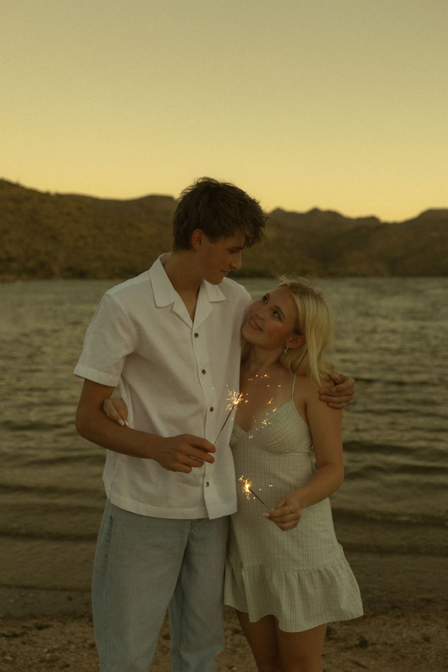 Young couple holding sparklers on a beach at sunset with mountains in the background.
