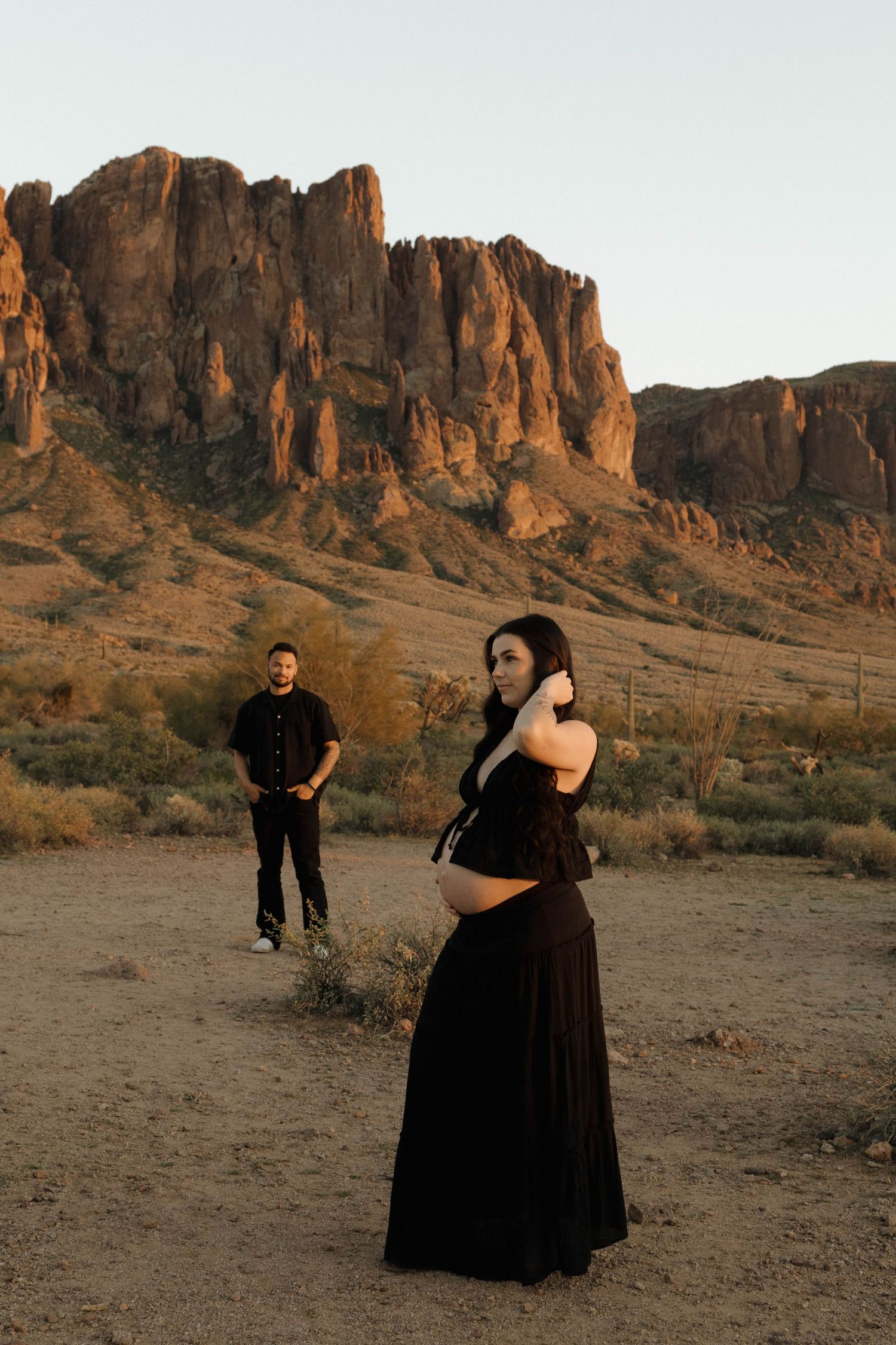 A couple in black attire dances and poses against dramatic desert rock formations at sunset.