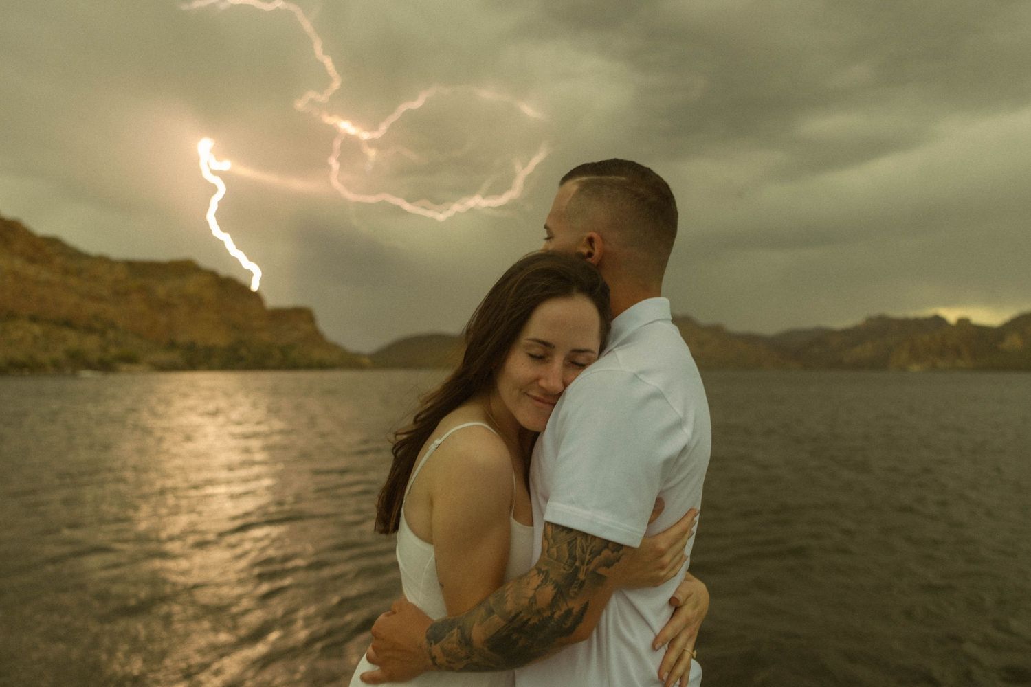 A couple embraces against a dramatic stormy sky with lightning and mountains in the background at Canyon Lake, Arizona.