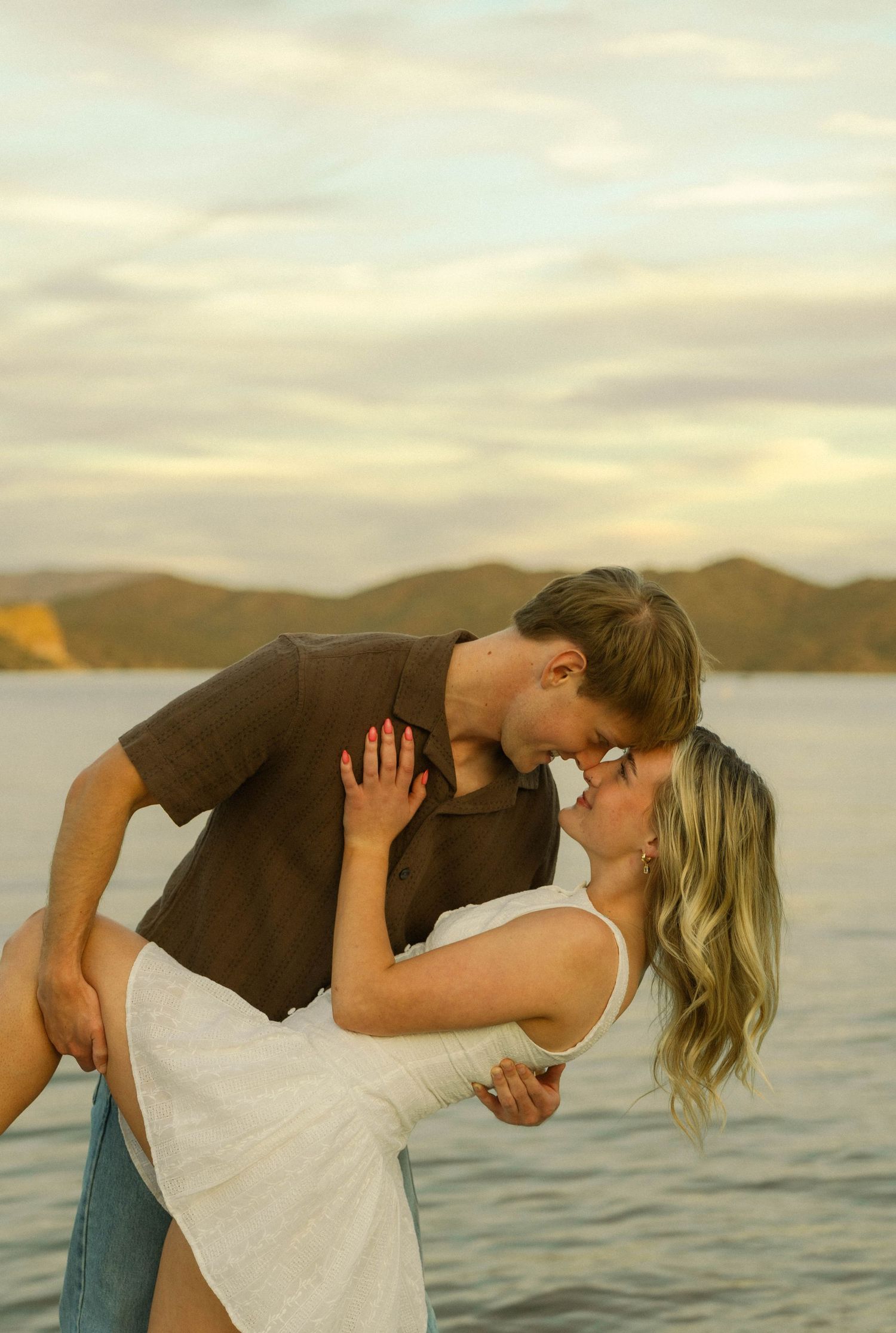 A couple shares a romantic moment by the water at sunset with mountains in the background.