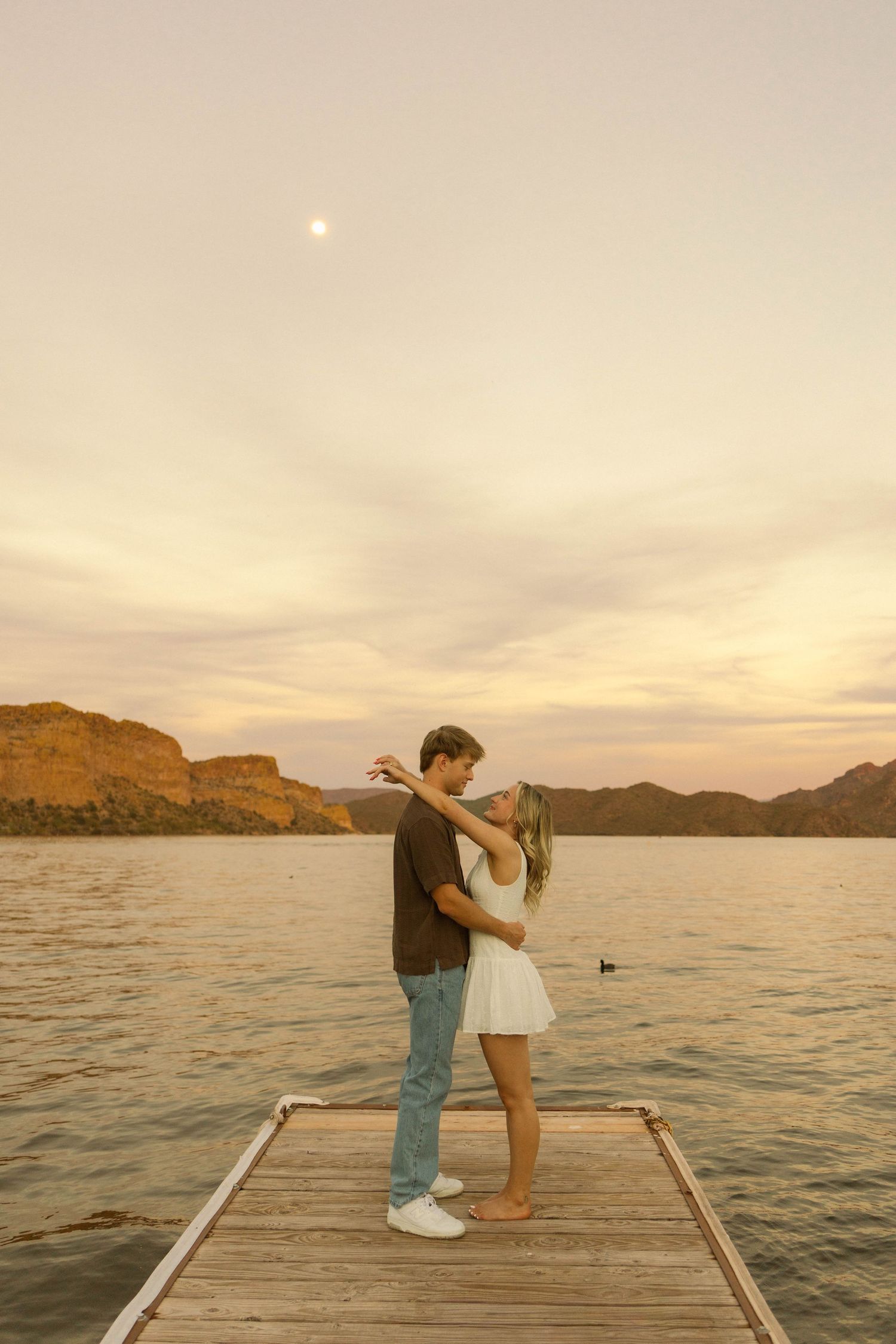 A couple embraces on a lakeside pier during golden hour with rugged mountains and calm waters in view.