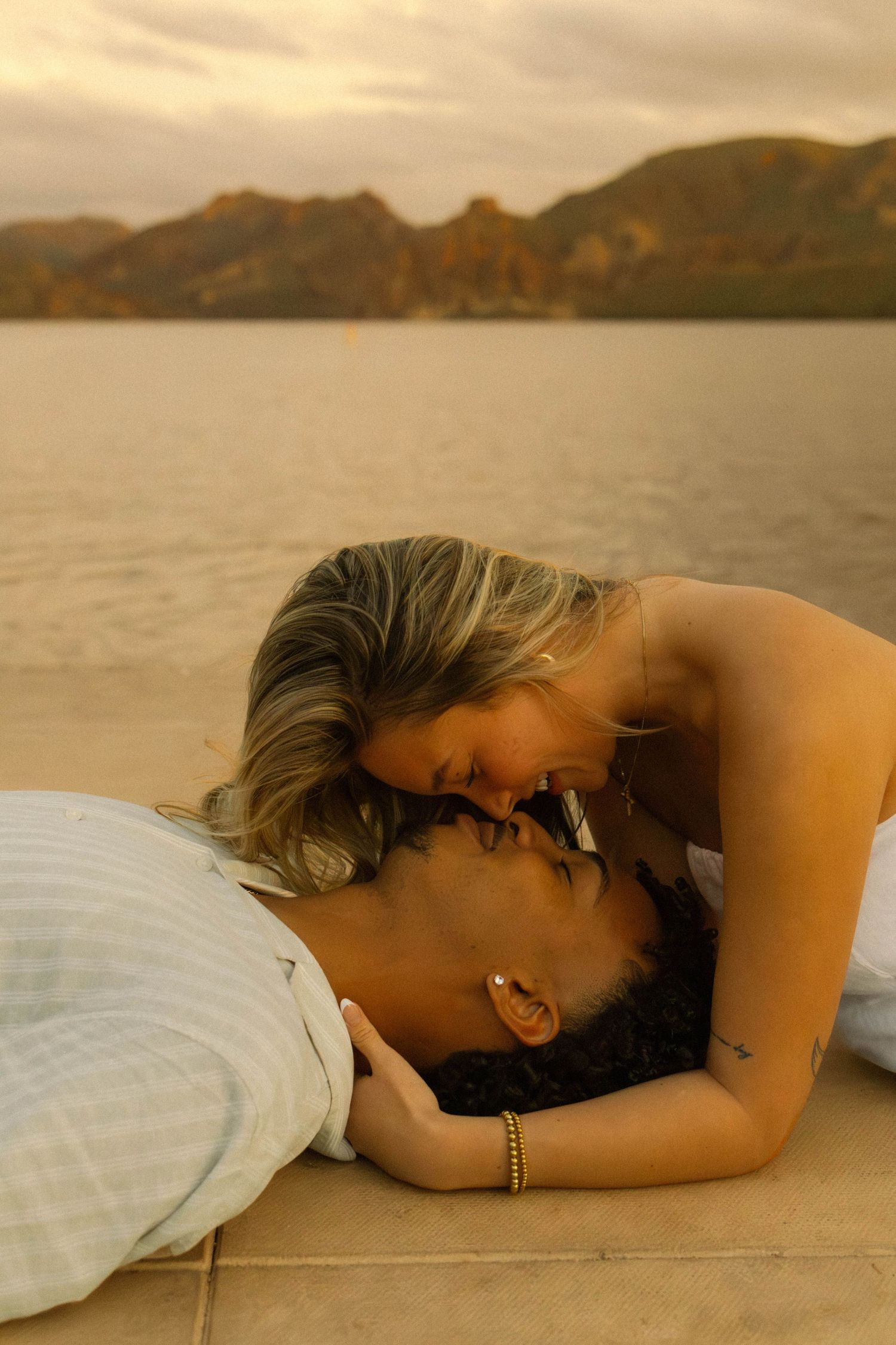 A person in a white dress lying down on a wooden dock at sunset with mountains in the distance.
