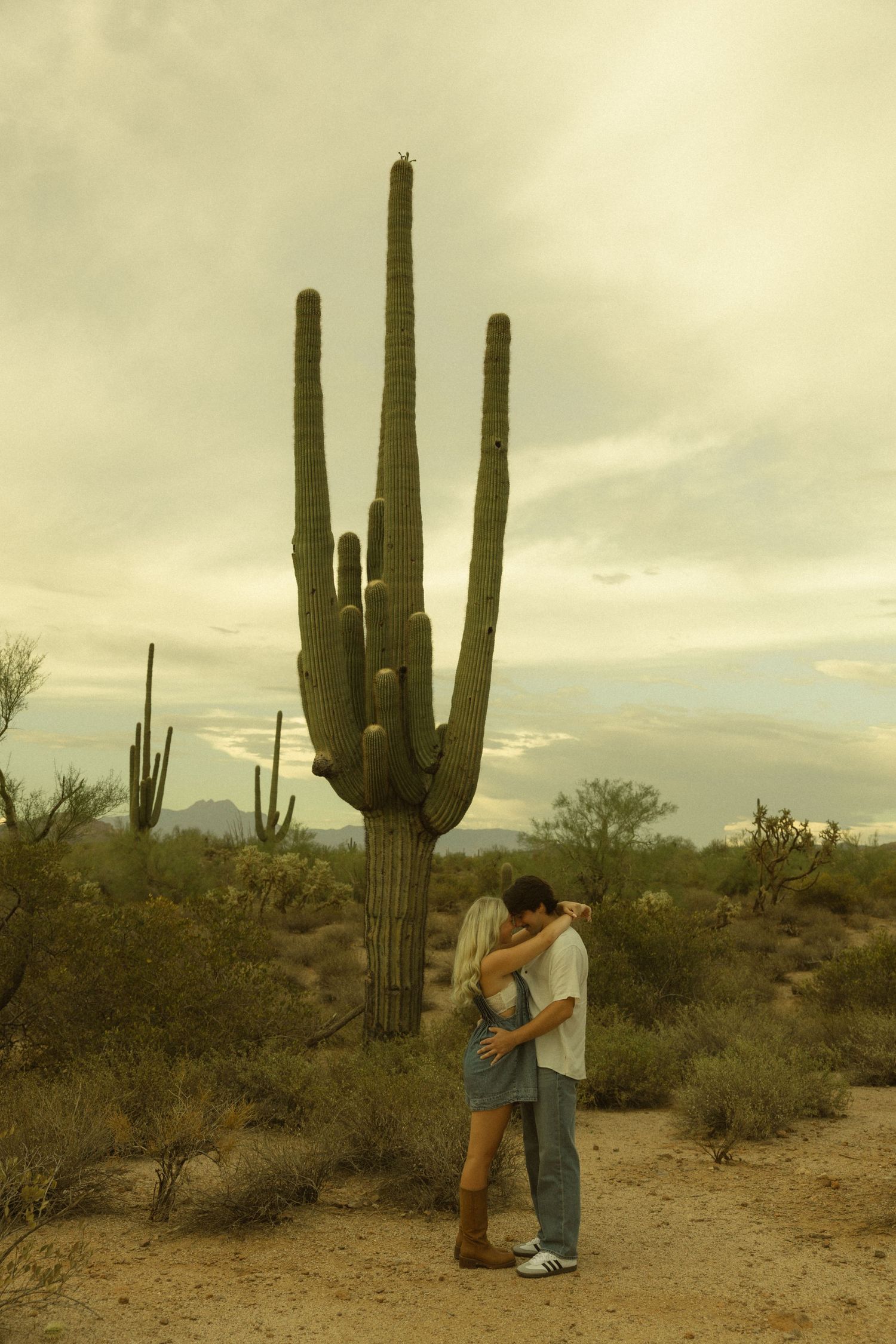 Couple embracing romantically in front of a tall saguaro cactus in the Arizona desert under a cloudy sky.