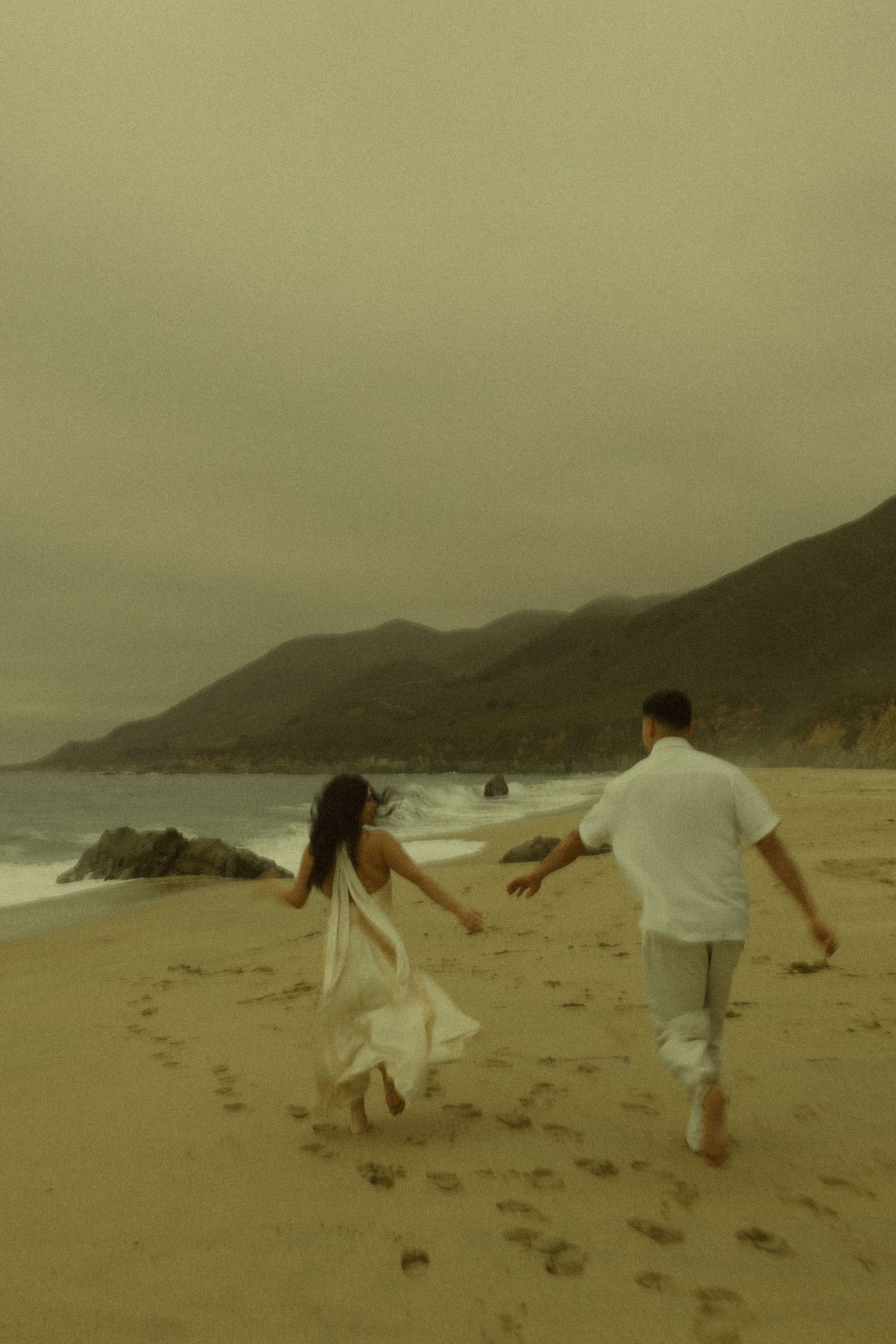 A couple in white clothing runs hand-in-hand along a sandy beach with misty mountains in the background.
