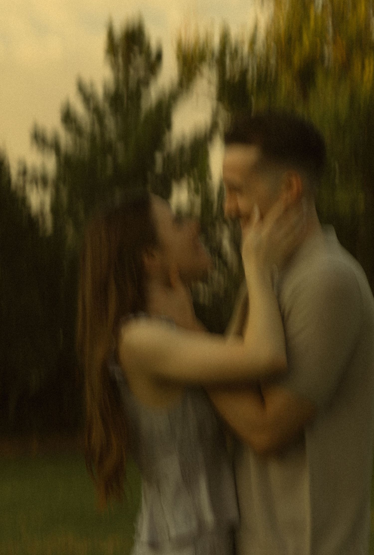 Blurry romantic couple embracing outdoors, woman touching man's face, surrounded by lush green trees.