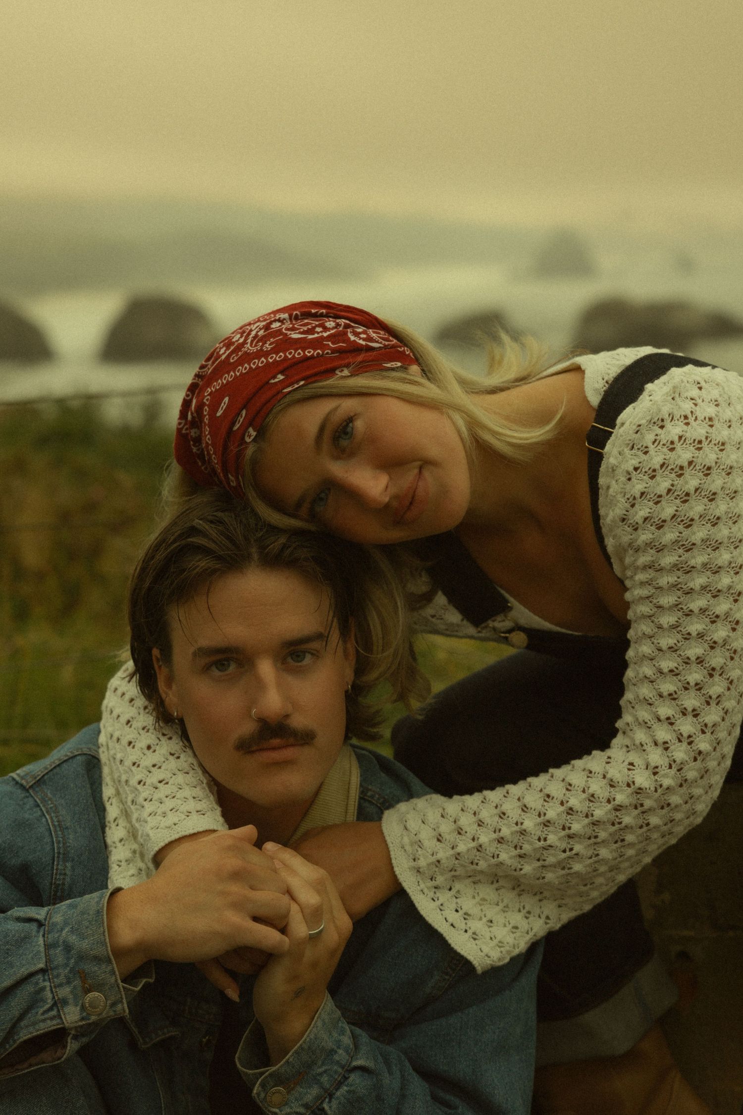 A young couple embraces outdoors near a misty coastal landscape, the woman wearing a red bandana resting her head on his shoulder.