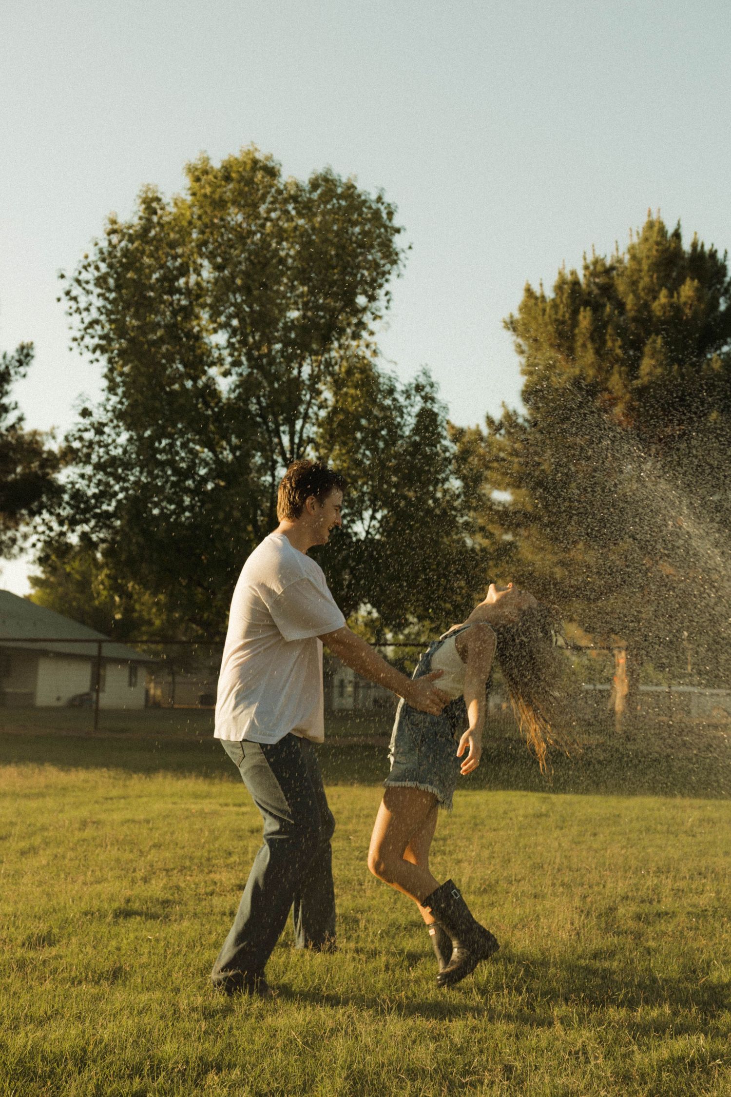 Couple dancing and spinning in a sunlit park, water droplets flying, surrounded by lush green trees.