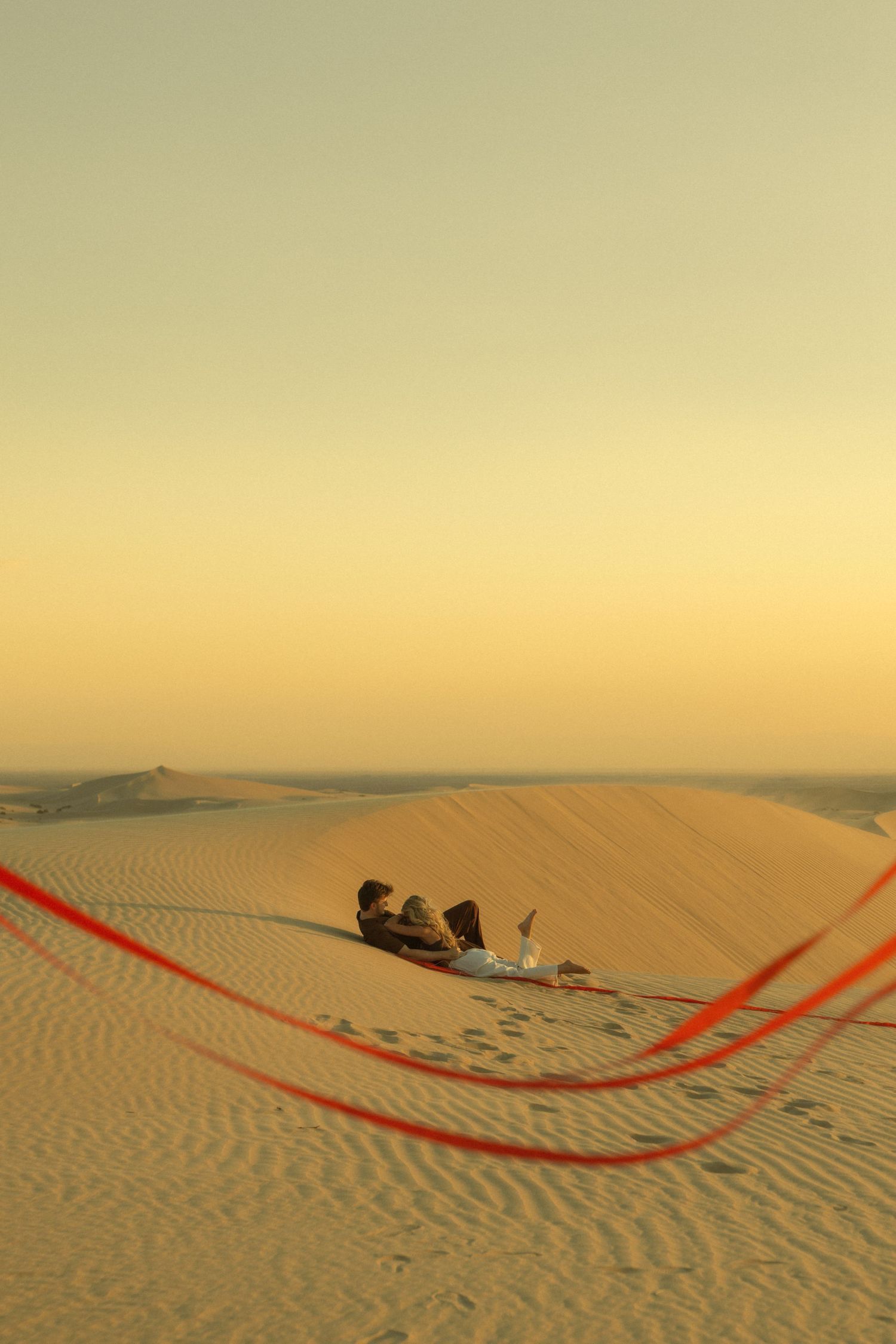 Couple relaxing on sand dunes at sunset with red kite string in foreground.