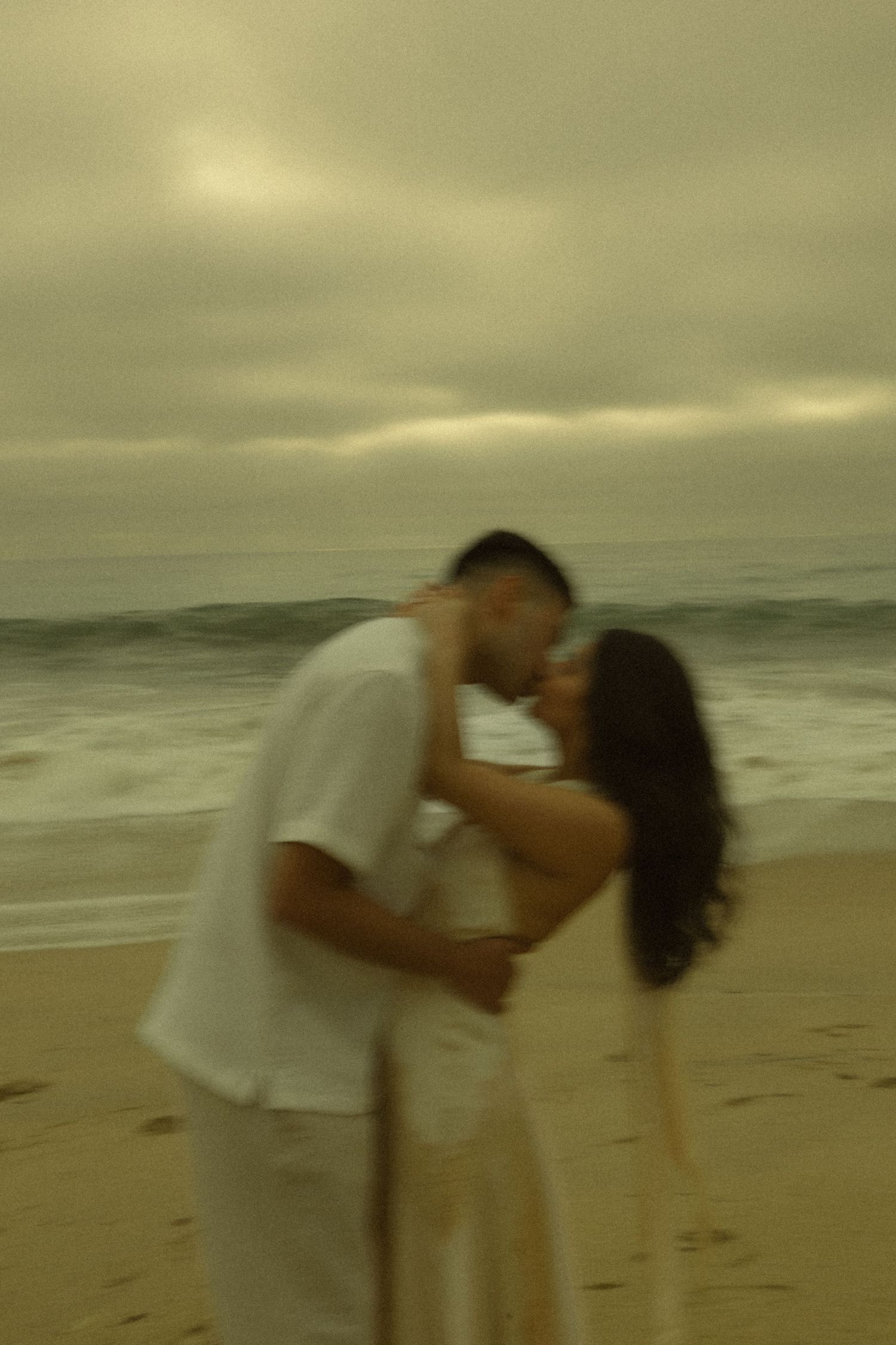 Couple sharing a romantic kiss on a sandy beach with ocean waves and cloudy sky behind them.