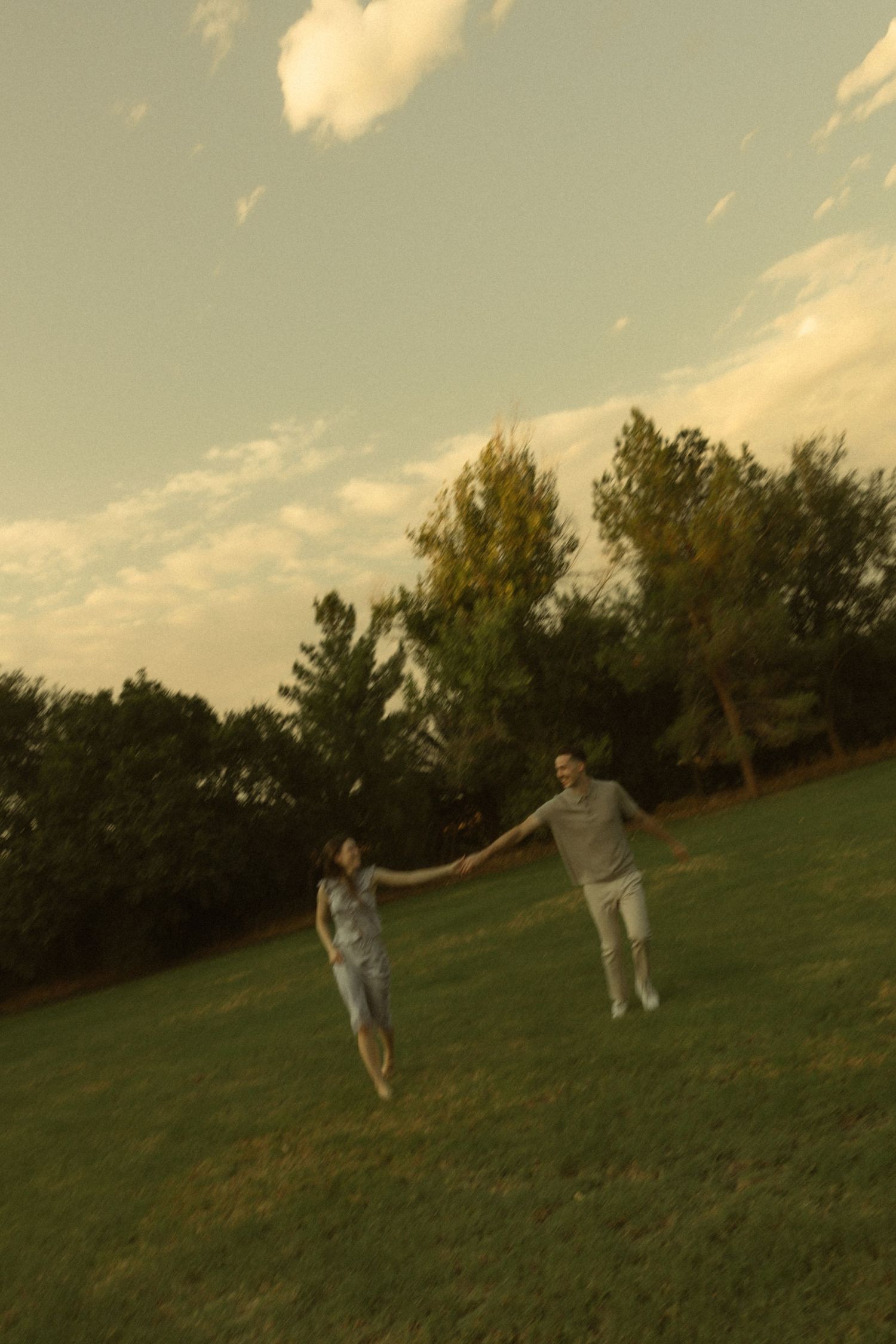 Couple holding hands and running together on green grass field with trees and cloudy sky at sunset.