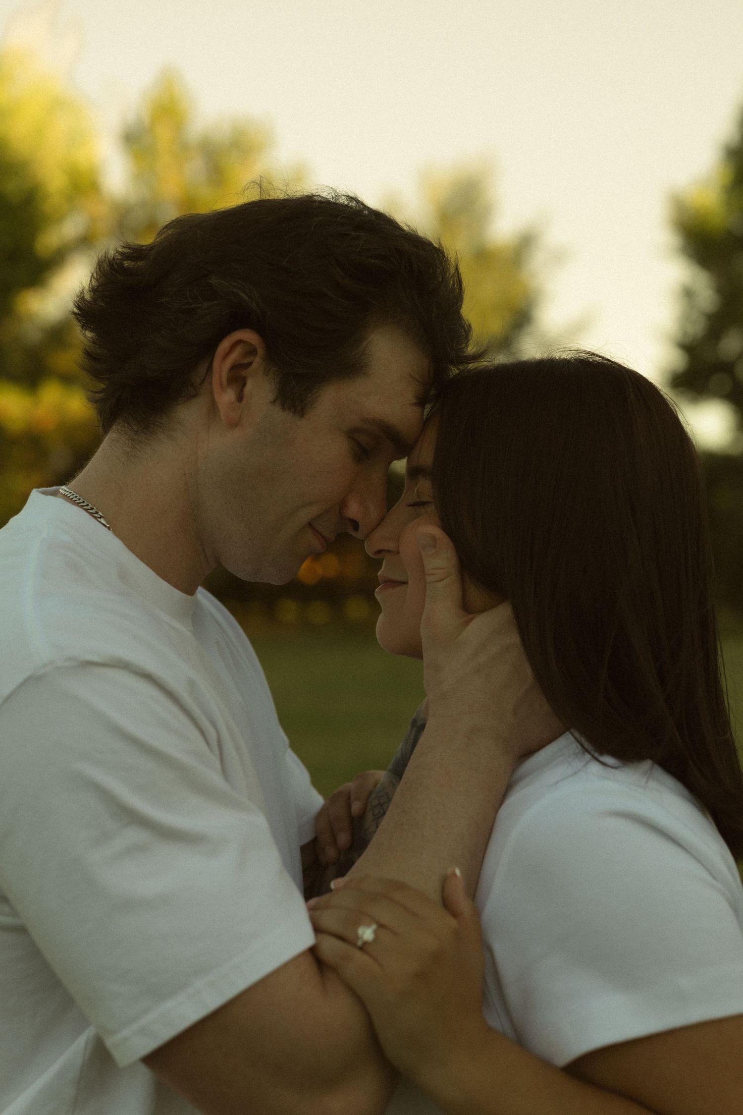 Couple in matching white t-shirts share a romantic forehead touch outdoors at sunset, woman wearing engagement ring.