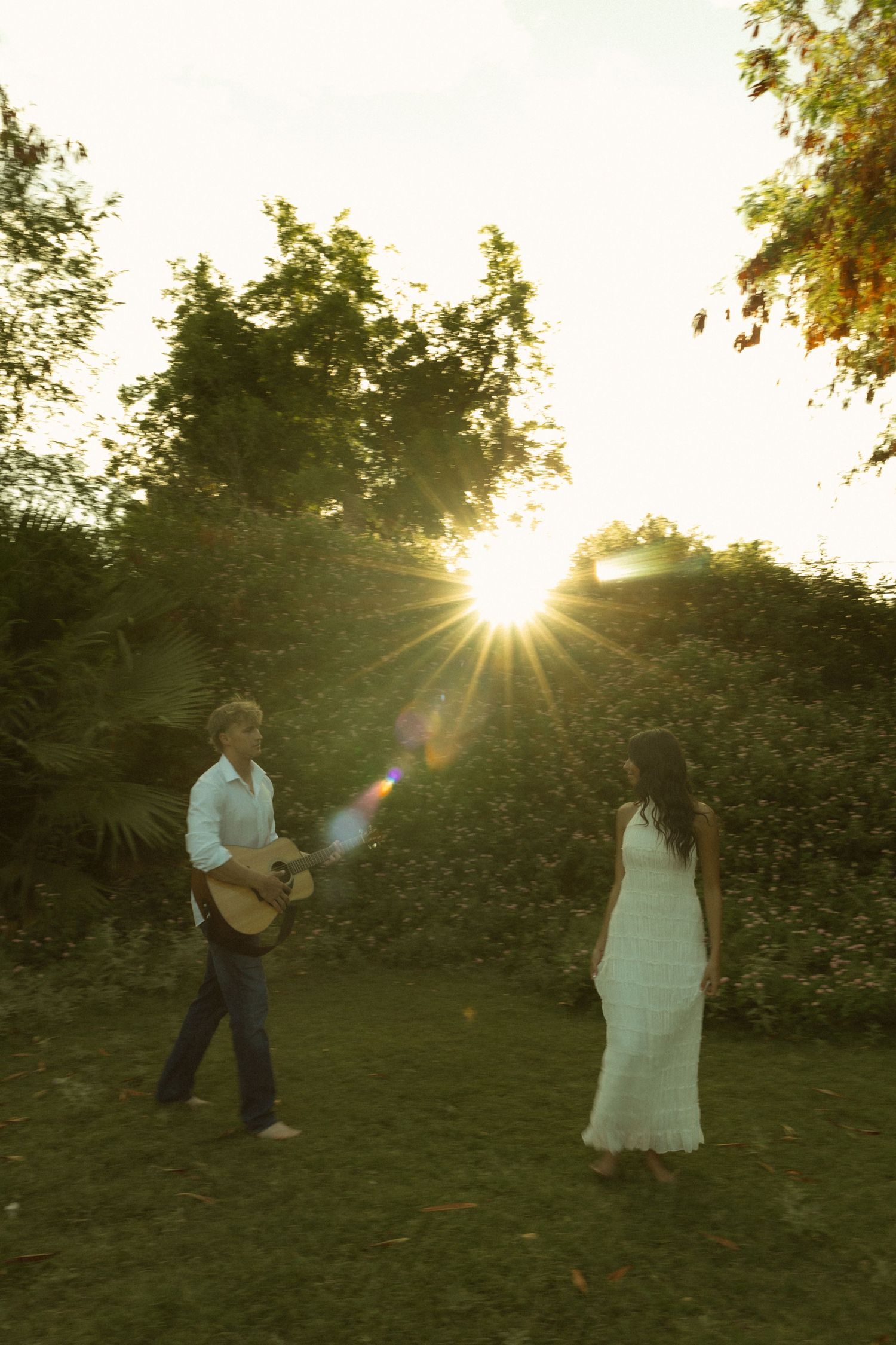 Couple standing in garden at sunset with sun flare, man serenading woman with acoustic guitar.