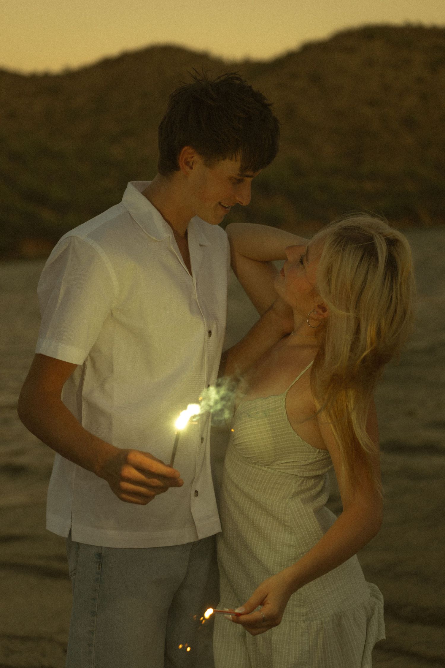 Young couple holding sparklers on a beach at dusk, smiling at each other in a romantic sunset setting.