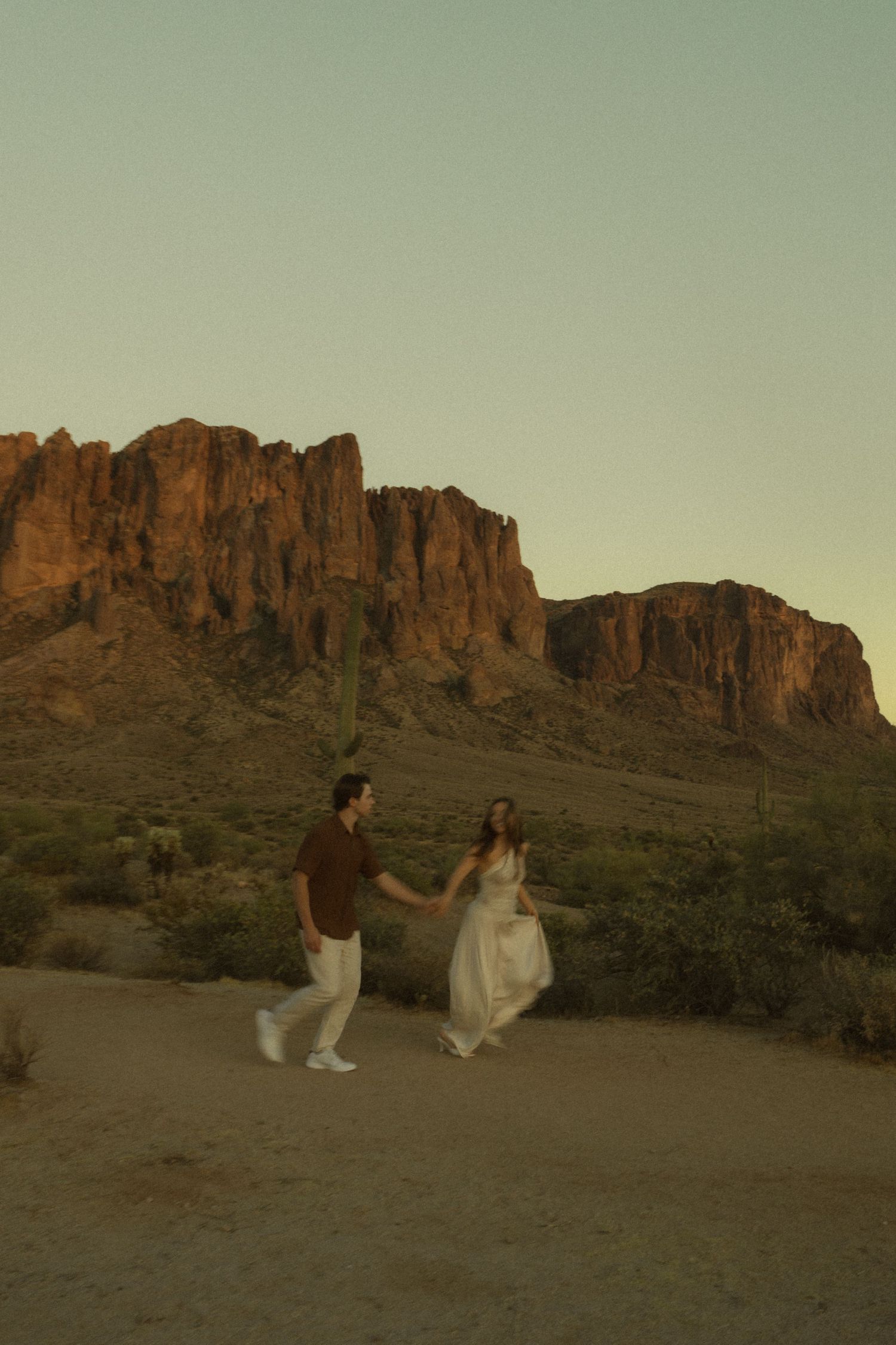 Couple holding hands walking in desert landscape with Superstition Mountains at golden hour sunset in Arizona.