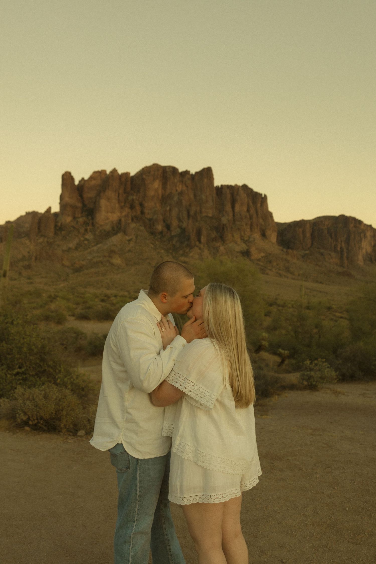 Couple kissing in white outfits with Superstition Mountains at golden hour in Arizona desert.