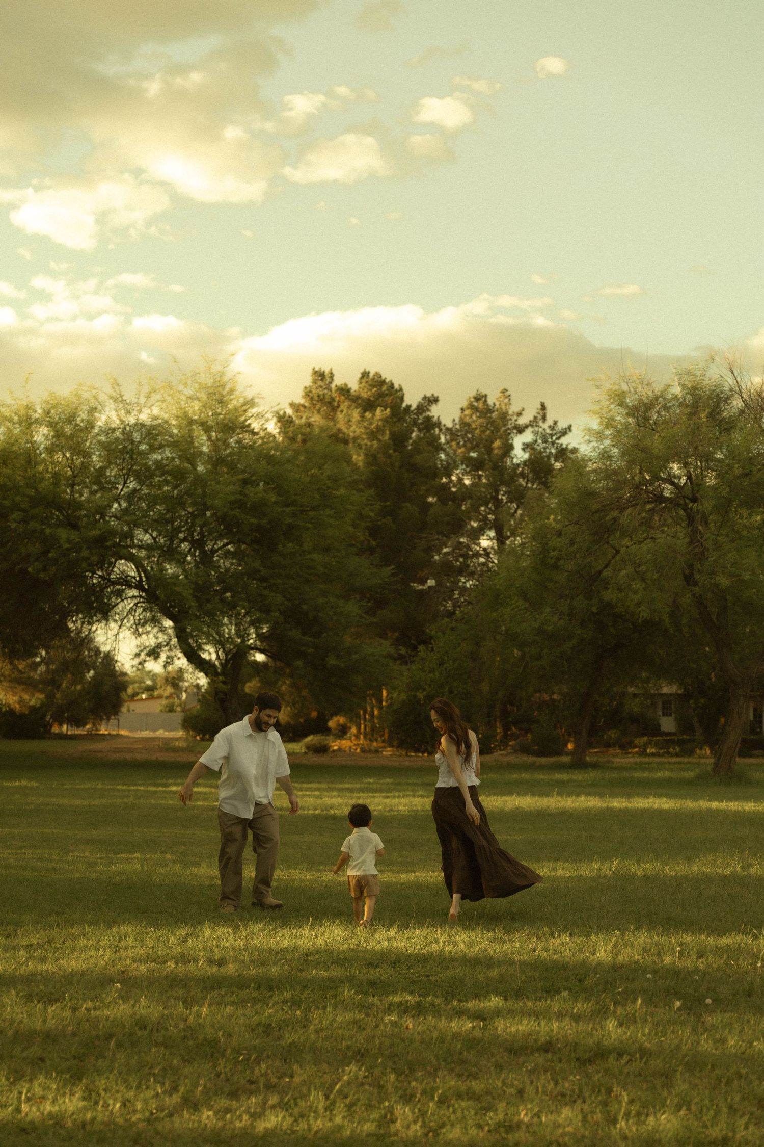 A family of three walks hand-in-hand across a lush green field at sunset with trees in the background.