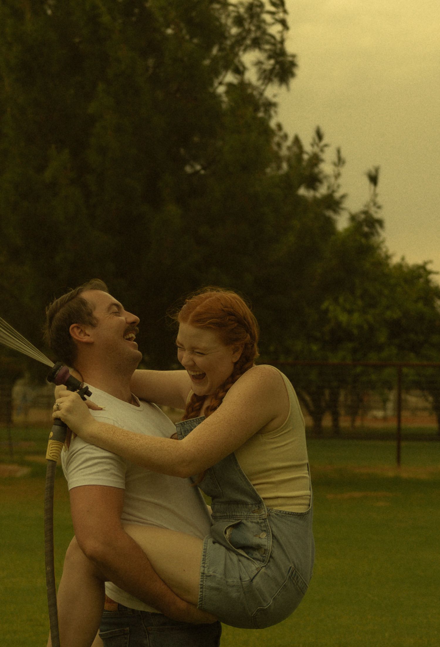 Couple laughing outdoors, man holding woman while spraying garden hose in lush green park setting.