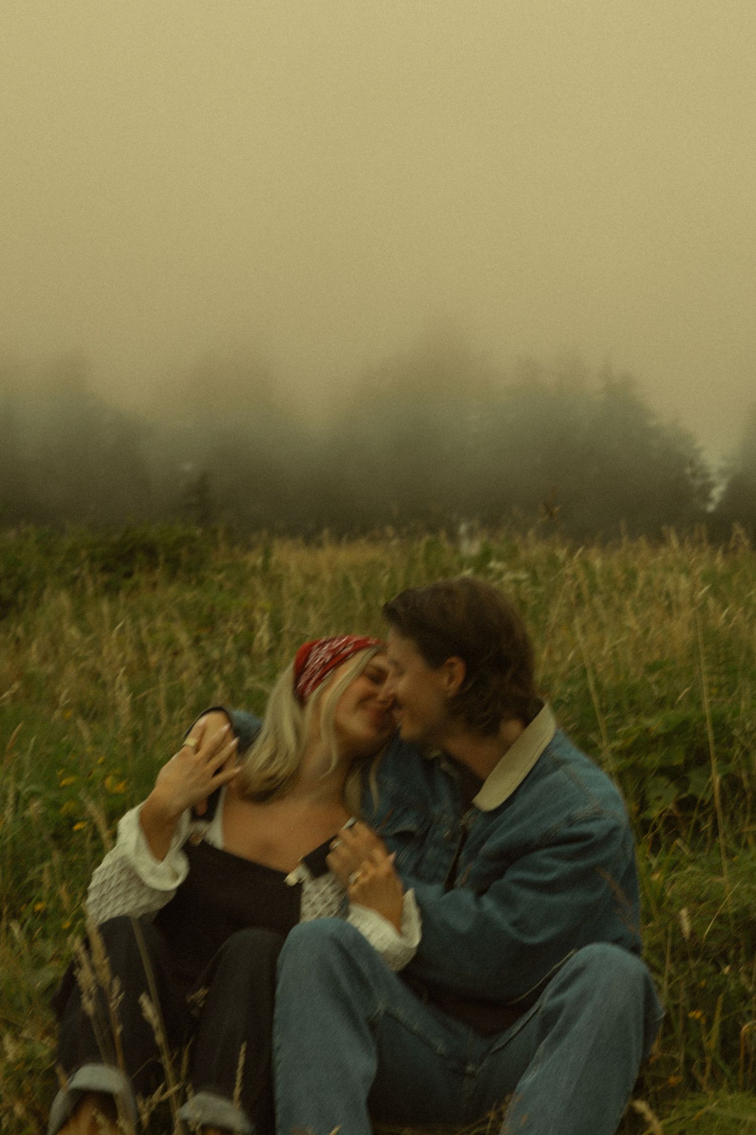Couple sharing a tender kiss in a misty meadow, woman in red bandana and white sweater, man in denim jacket.