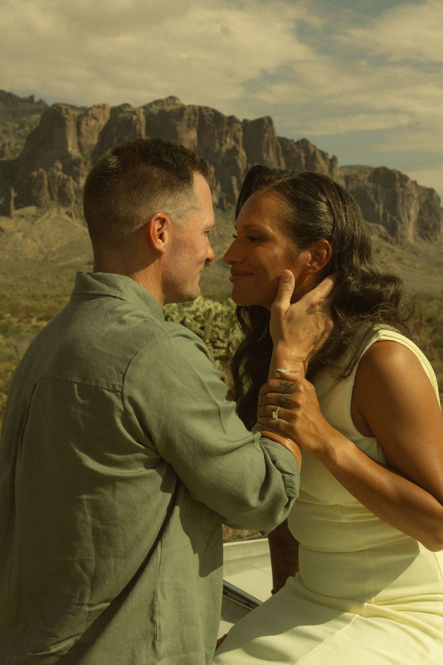 Couple sharing a romantic moment in the desert with rugged mountain backdrop under a cloudy sky.