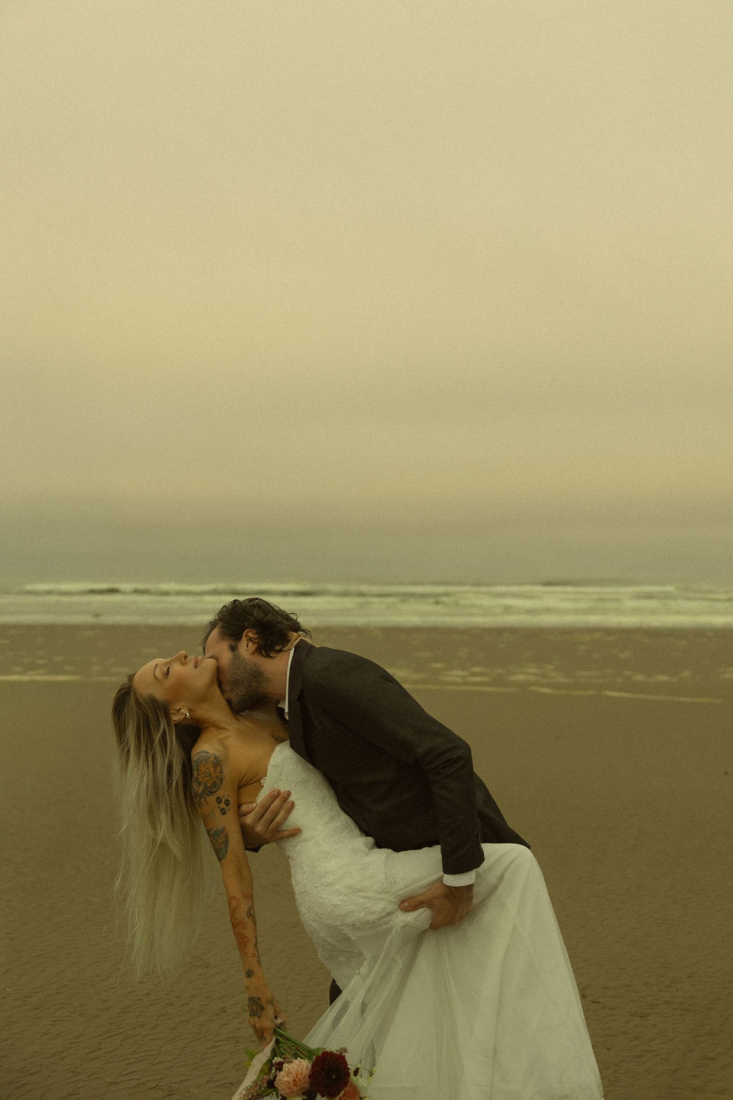 Bride and groom share a romantic dip kiss on a misty beach during their oceanside wedding ceremony.
