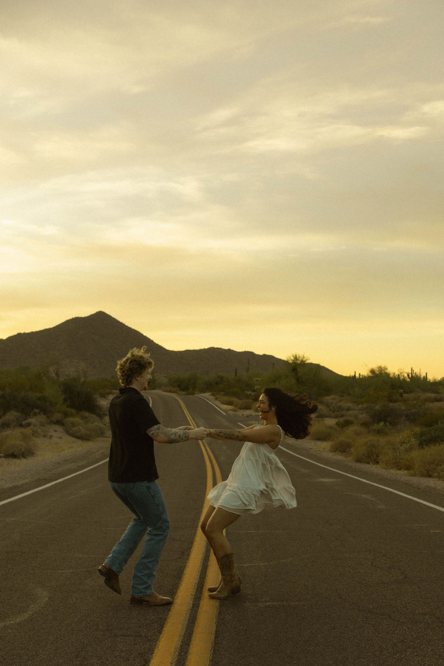 Couple dancing together on a desert road at sunset with mountains in the background.