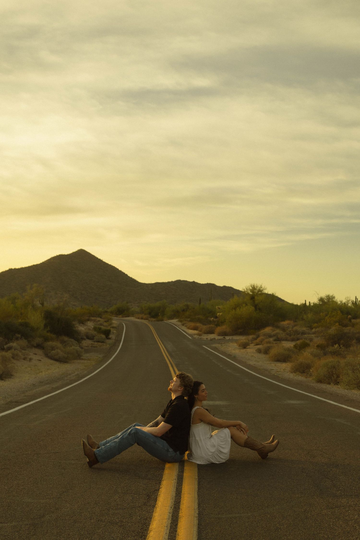 Couple sitting back-to-back on desert road centerline at dusk, surrounded by arid landscape and dramatic sky.