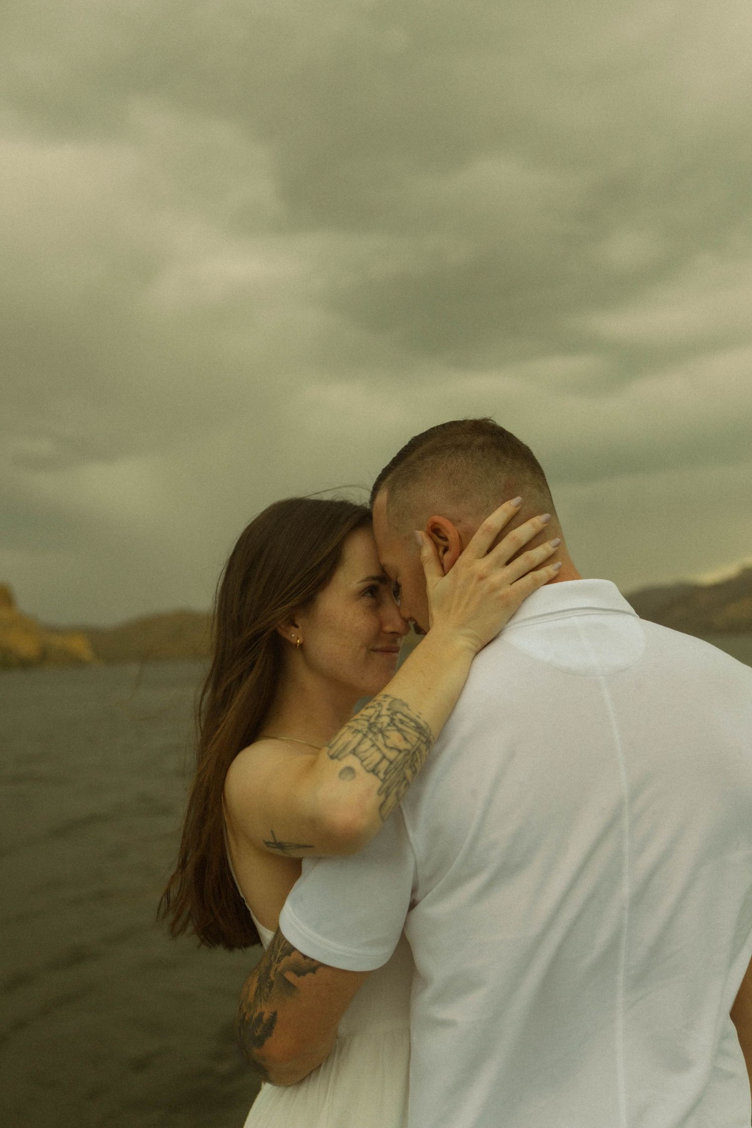 A tender embrace between a couple wearing white outfits near a scenic waterfront with mountains in the distance.
