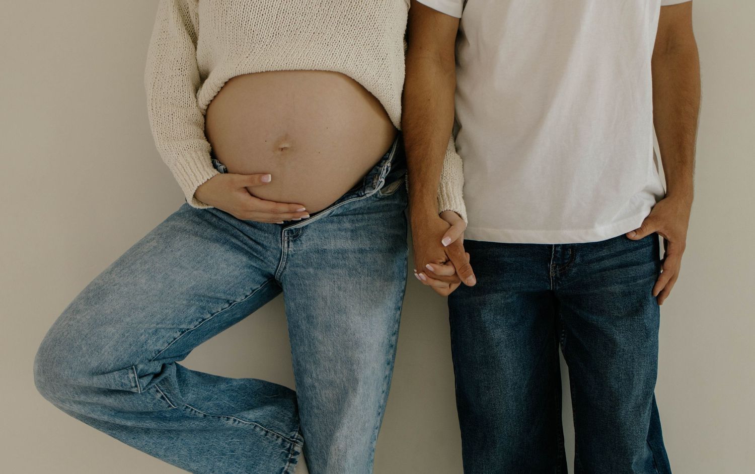 Cropped view of an expecting couple holding hands in casual jeans and neutral tops against a white wall.