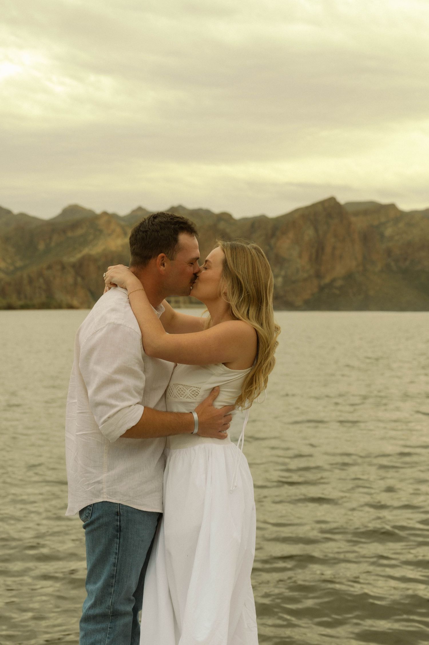 A romantic couple shares a kiss at sunset by a lake with dramatic mountain peaks in the background.