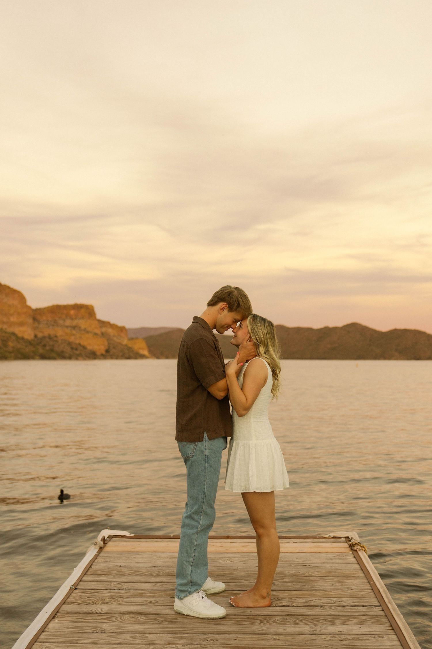 A couple shares a romantic moment on a wooden dock at sunset overlooking a serene lake and mountains.