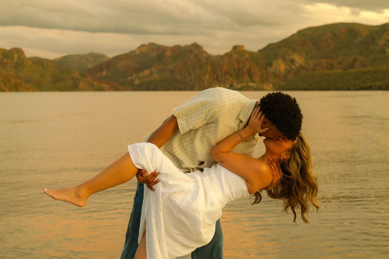 Two people in white clothing dance playfully by a lake at sunset.