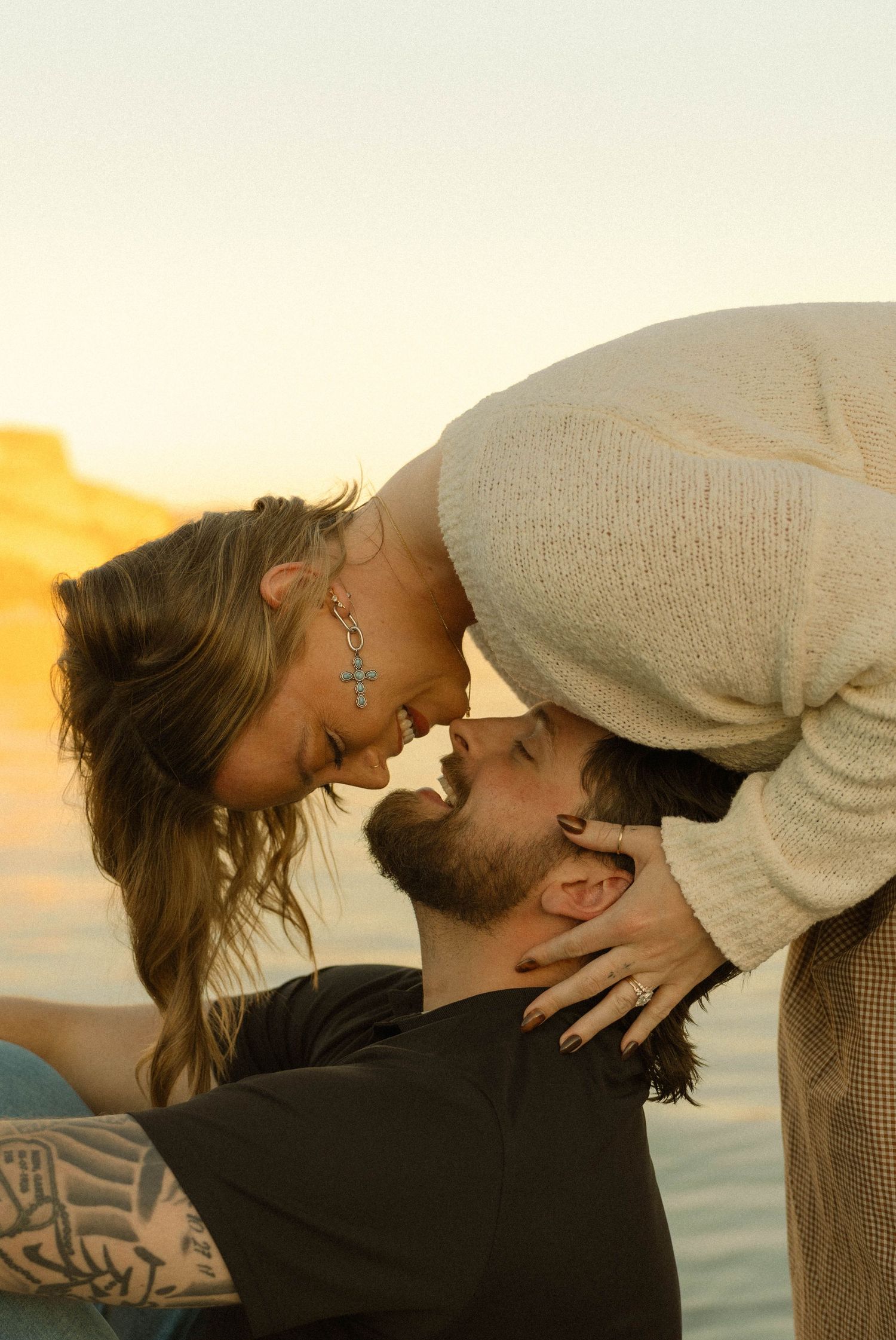 Romantic couple sharing an intimate moment at sunset by the water with tattoos and casual attire.