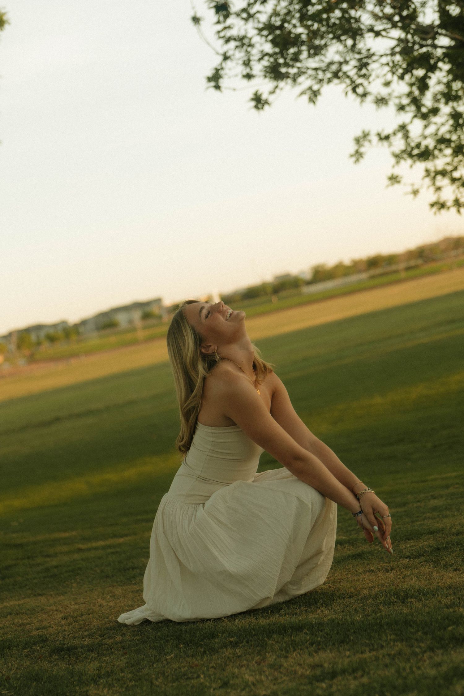 A joyful woman in a white dress kneels on green grass, laughing with arms back at golden sunset.