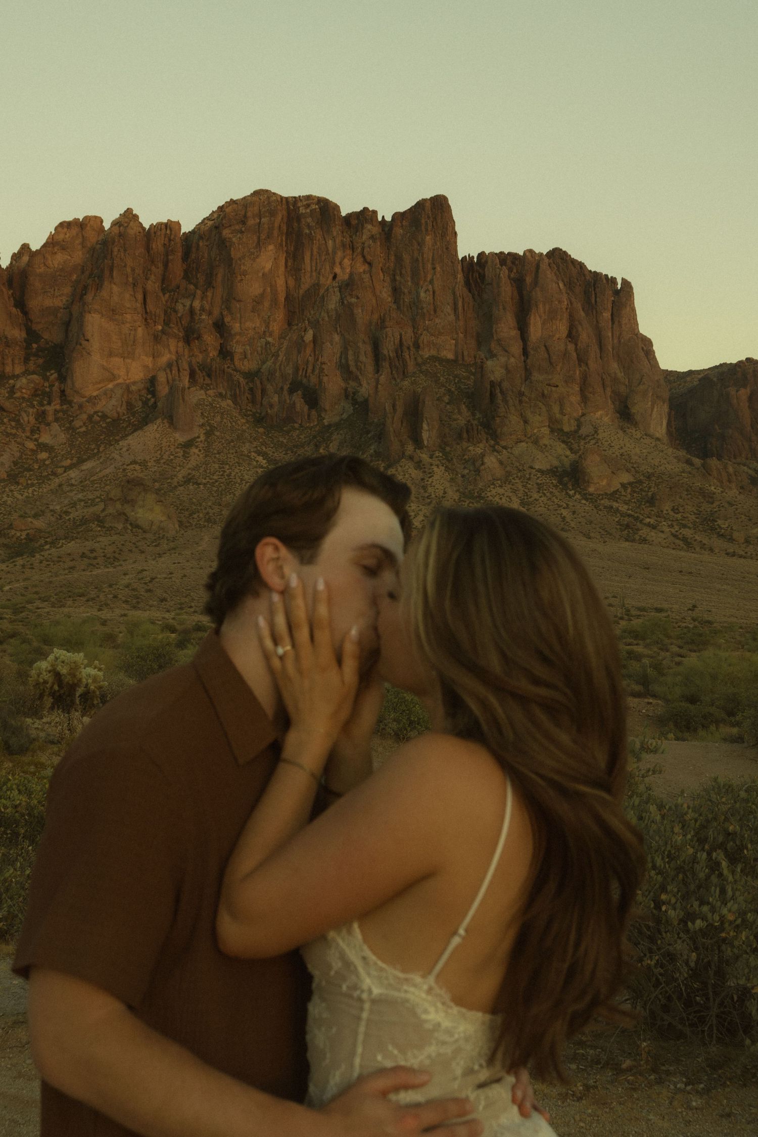 Couple sharing a kiss with dramatic red rock mountains in the Arizona desert at sunset.