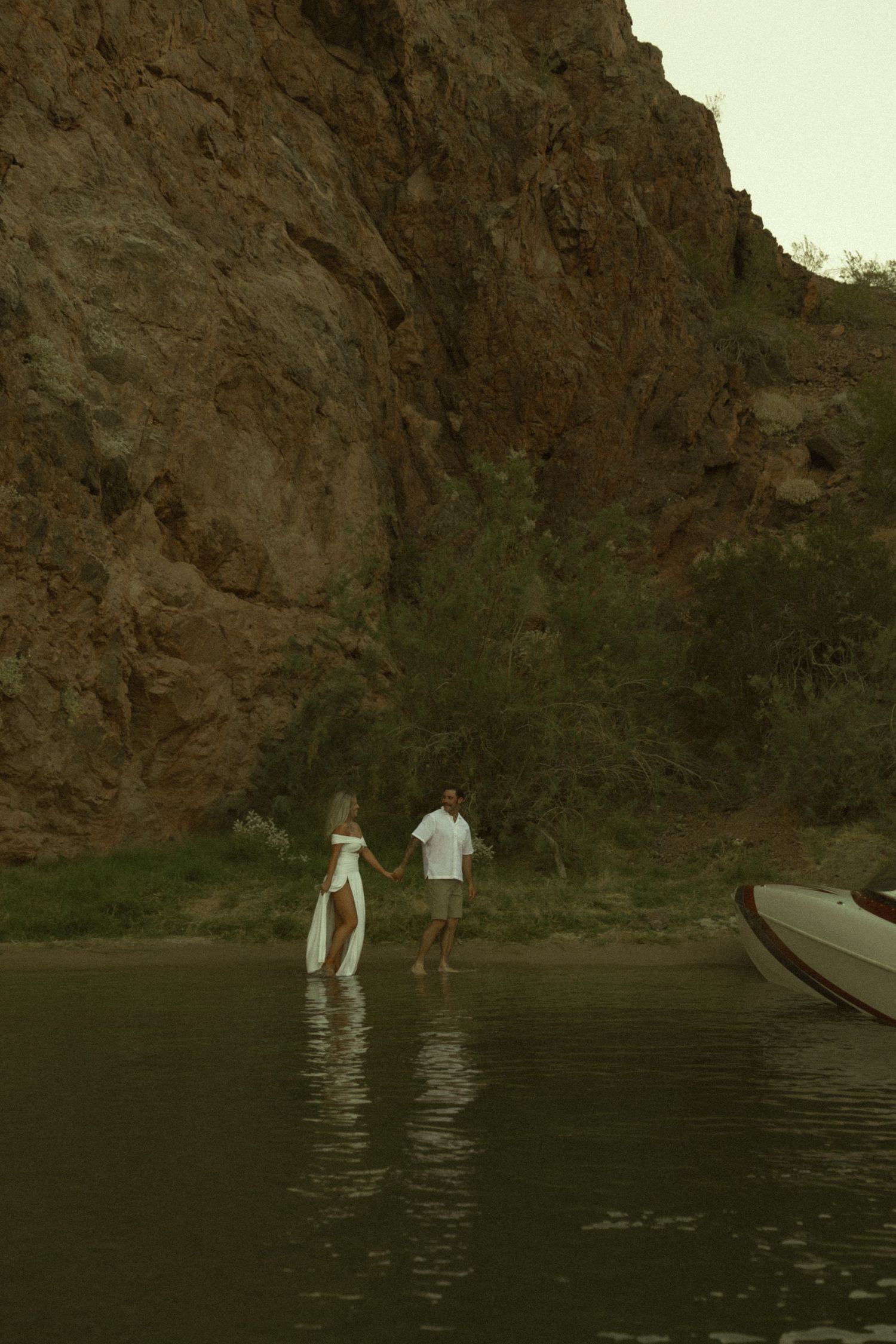Couple holding hands wading through shallow water near rocky canyon cliffs beside a moored boat.