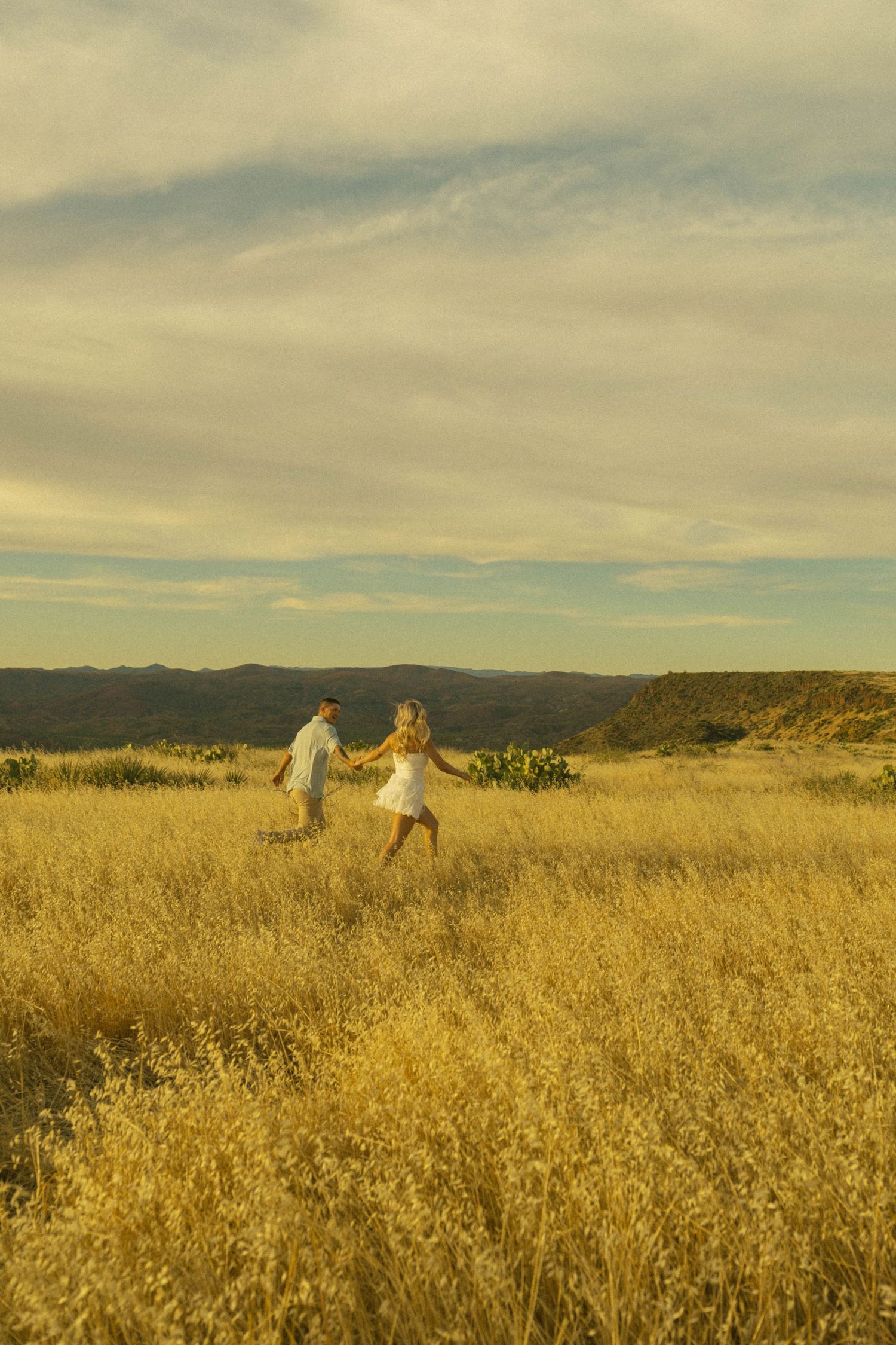 Couple holding hands walking through golden grass field with mountain backdrop under cloudy sky at sunset.