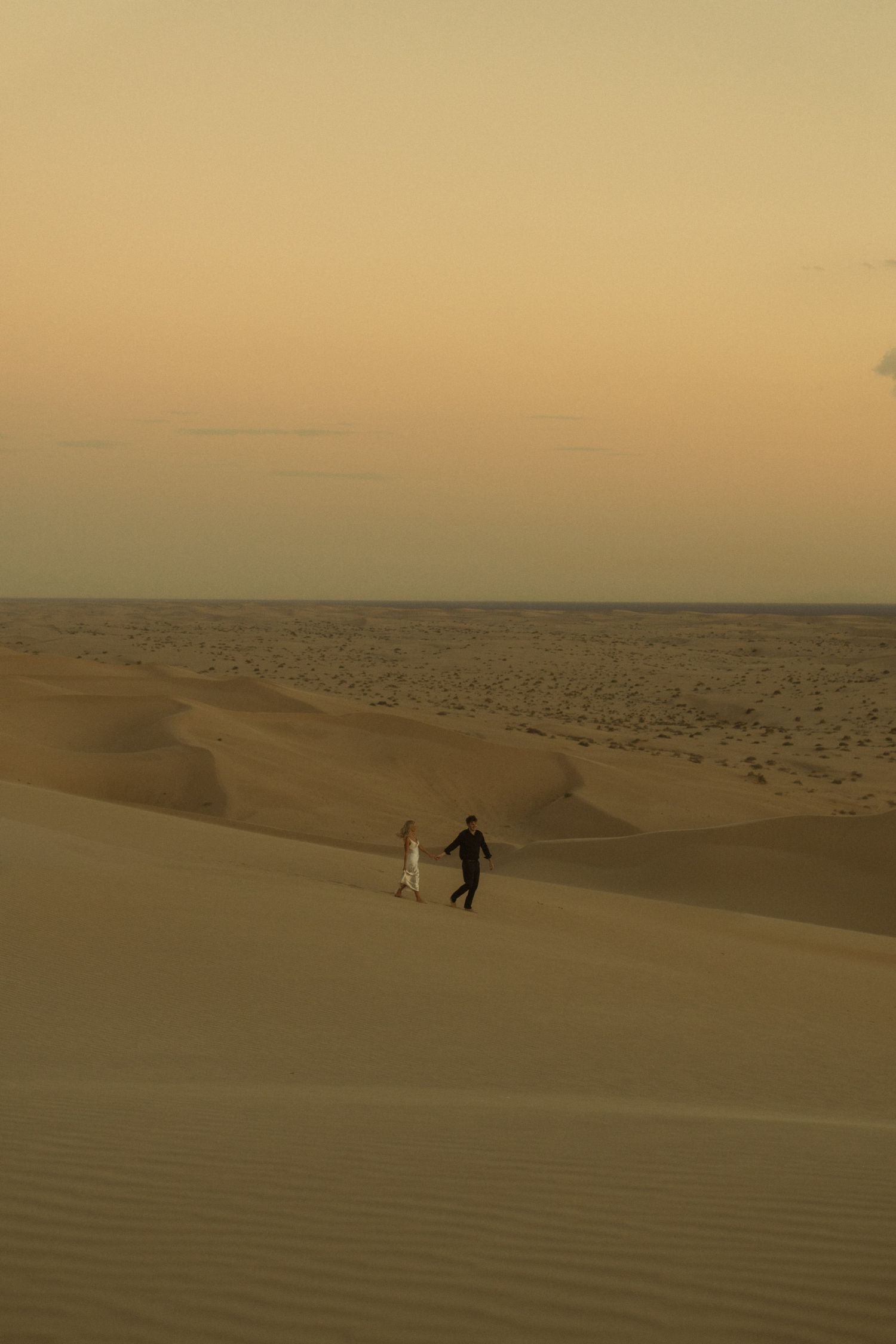 Couple holding hands walking across expansive golden sand dunes at desert sunset.